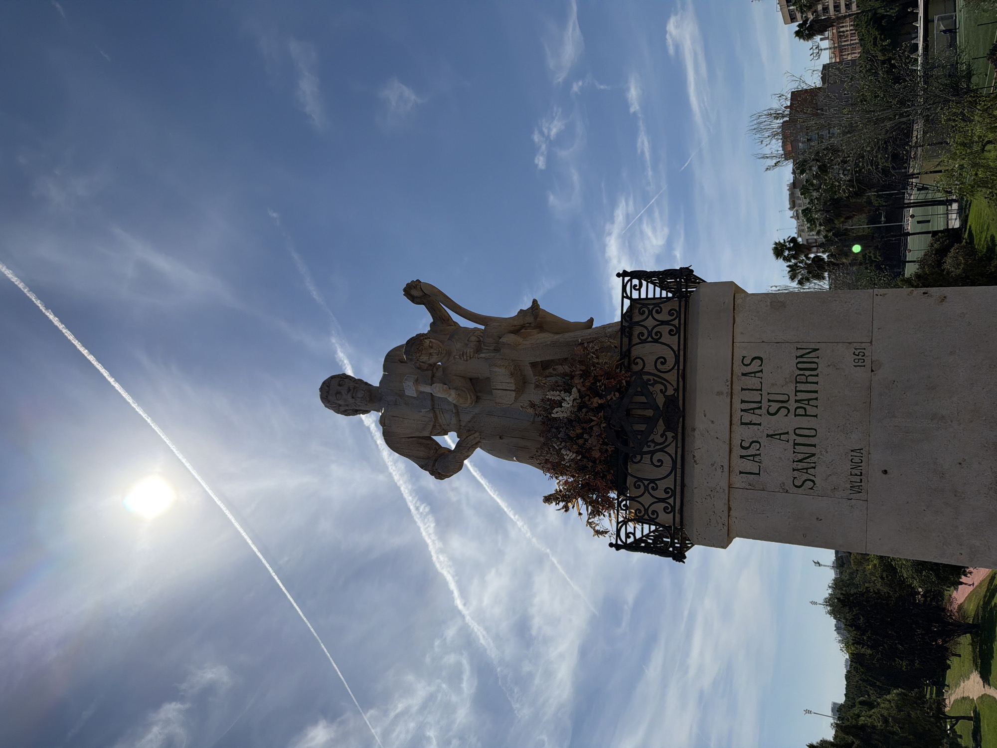 The image depicts a statue in València, Spain. The statue, made of light-colored stone, depicts a man standing, possibly a religious figure given the inscription, with two smaller figures around him. The statue is mounted on a stone pedestal with a wrought-iron railing. The pedestal is inscribed with the words "LAS FALLAS A SU SANTO PATRON VALENCIA 1951". The background shows a blue sky with wispy clouds, and buildings and greenery in the distance. The overall scene appears to be a public park or square.