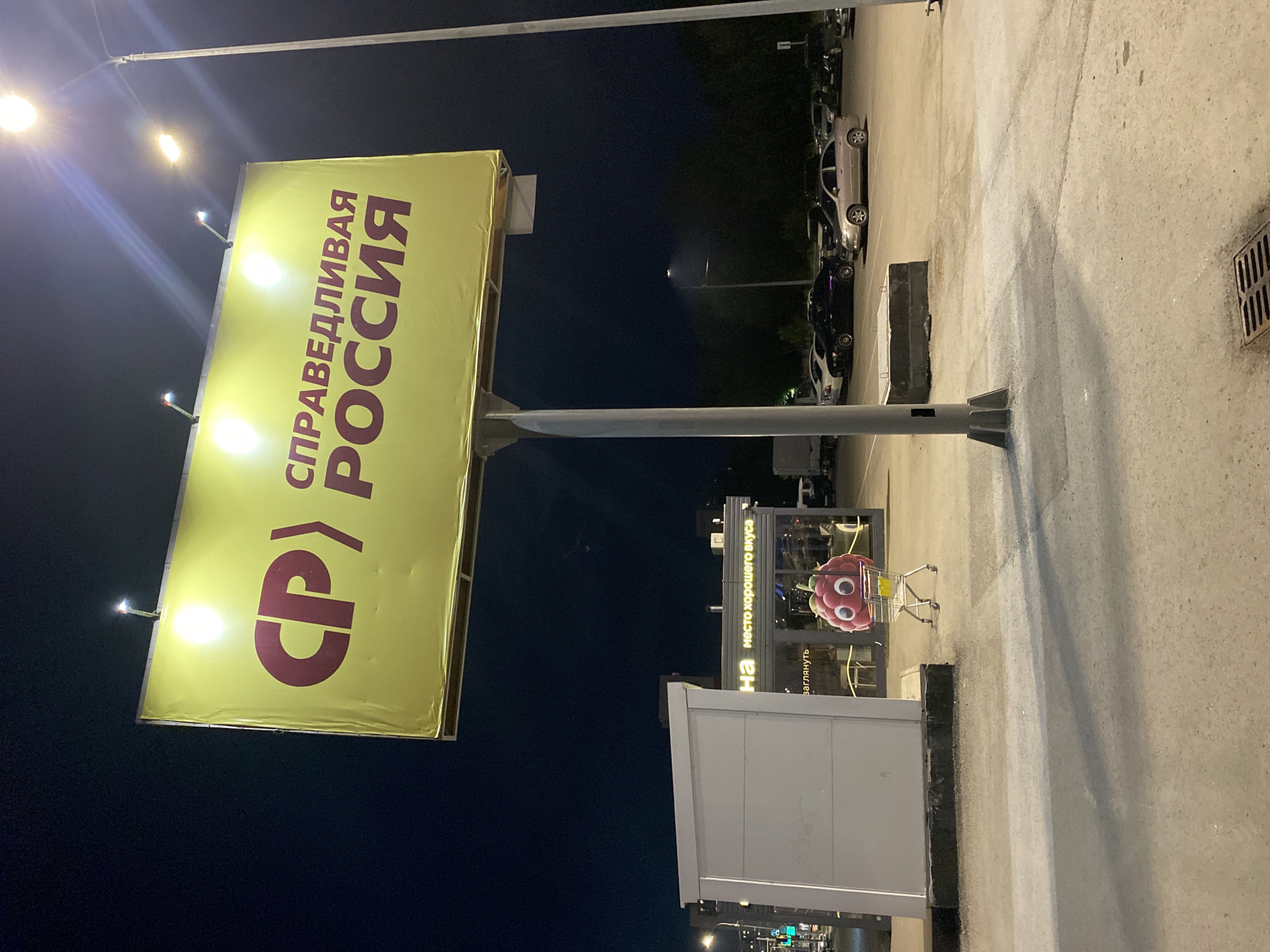 A brightly lit yellow billboard with red text and a stylized logo stands prominently in a nighttime outdoor scene in Novokuybyshevsk, Russia. The billboard displays the Cyrillic text "СПРАВЕДЛИВАЯ РОССИЯ" (Spravedlivaya Rossiya, meaning "A Just Russia"), a political party. Below and to the left of the billboard, a small, white structure with a sign reads "НА МЕСТО ХОРОШЕГО ВКУСА" (Na Mesto Khoroshego Vkusa, roughly translating to "To the Place of Good Taste"). Next to this building, a whimsical, cartoonish representation of a raspberry character with large eyes is positioned inside a shopping cart. The area appears to be a parking lot, with several cars parked in the background. Streetlights illuminate the scene, casting long shadows and highlighting the textures of the asphalt and concrete ground. The sky is dark, indicating it is nighttime.