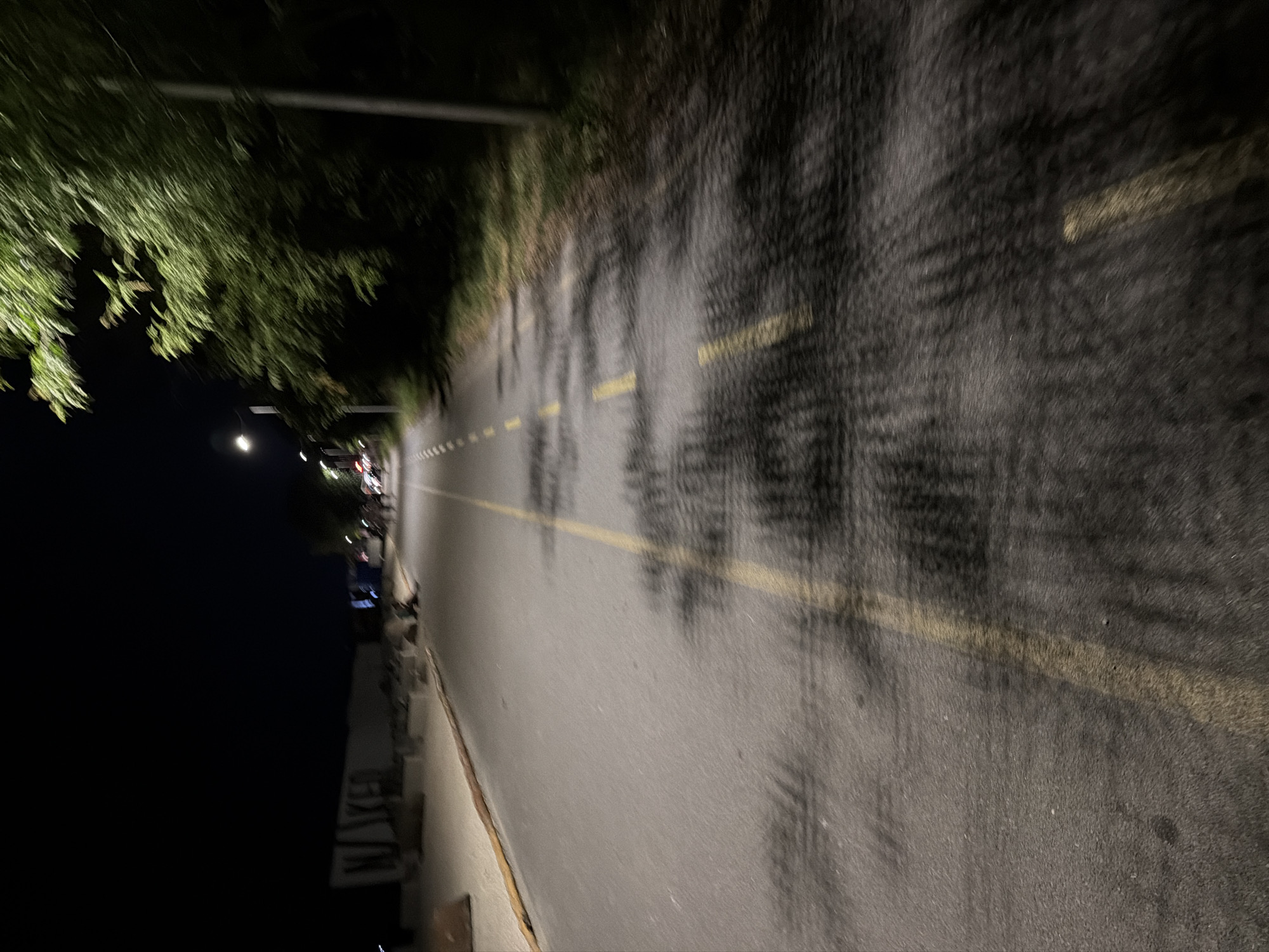 The image shows a road at night, likely a promenade or bike path, with yellow markings. On the left side of the road, there is a sandy area with a white structure featuring large lettering that appears to spell out "NAKED". Further down the road, there are streetlights and trees. The image has a blurred effect, suggesting movement. The location is identified as the northern beach area in Burgas, Bulgaria.
