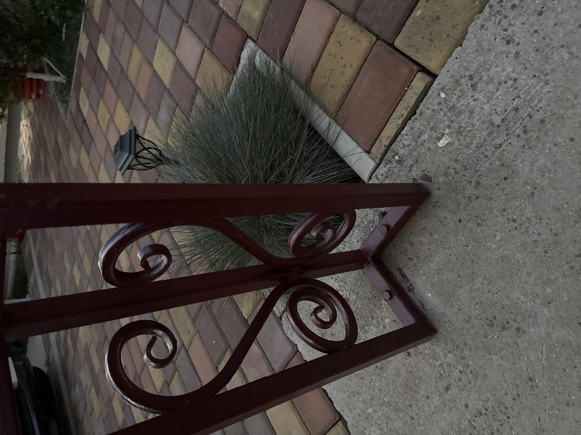 The image is a close-up, ground-level view of a dark red metal fence with decorative scrollwork in the foreground. Beyond the fence, a cobblestone pathway is visible, laid with rectangular pavers in shades of red, yellow, and brown. To the right of the pathway, a clump of dense, spiky green grass grows in a rectangular cutout. A small, black, square-topped garden light with a decorative wire base is partially obscured by the grass. In the upper left corner, the rear tire and lower body of a dark-colored vehicle are visible. The environment appears to be an outdoor setting, possibly a courtyard or residential area. The scene is static and shows no human activity.