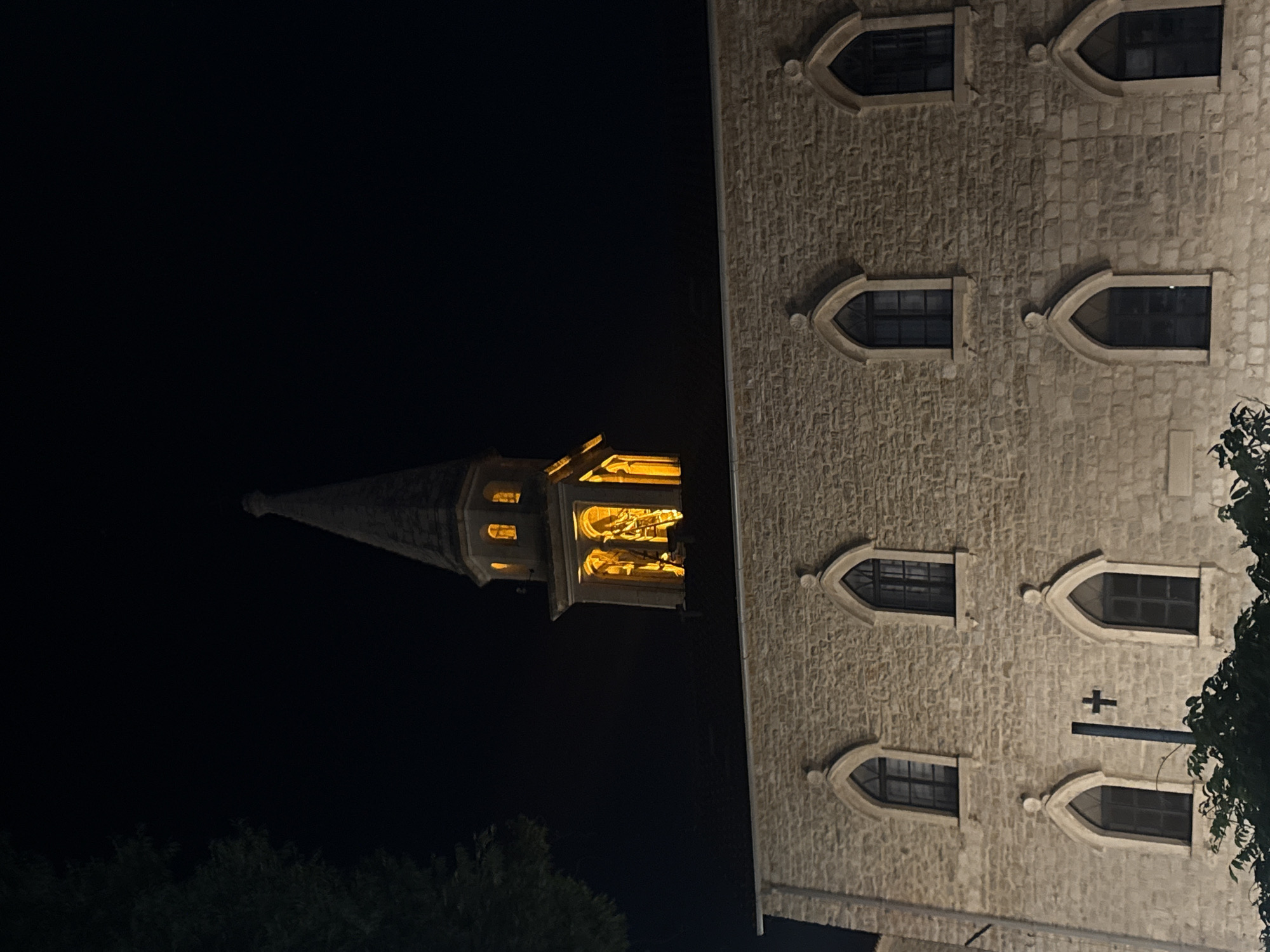 The image shows a stone building with a tall, pointed spire illuminated by yellow lights at night. The building has multiple arched windows. A dark tree is visible on the left side of the frame. This building appears to be the Church of Saint John the Baptist in the Old Town of Budva, Montenegro, a prominent landmark often photographed at night due to its illuminated spire.
