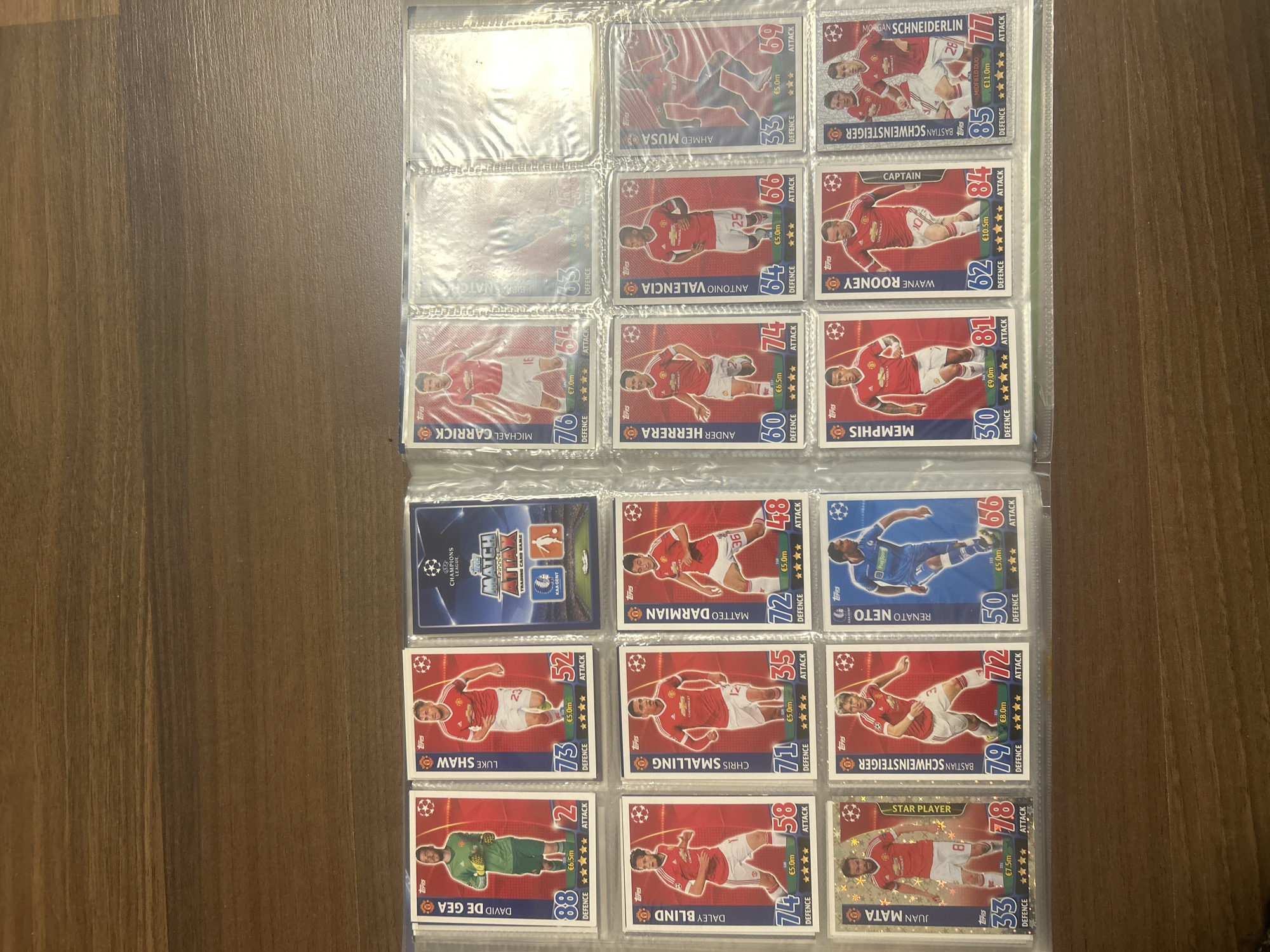 The image shows a collection of football trading cards in a plastic sleeve binder. The cards are from a "Match Attax" Champions League series, featuring various football players. Most of the cards showcase players from Manchester United in their red kits, with occasional cards from other teams, such as one featuring Renato Neto in a blue kit. Each card displays a player's name, a photo, and various statistics related to attack and defense. Some notable player names visible include Wayne Rooney, Bastian Schweinsteiger, David De Gea, Juan Mata, and Luke Shaw. The cards are arranged in a grid-like formation within the plastic sleeves. The background is a brown, wooden surface. The location is not known to be Pleven, Bulgaria.