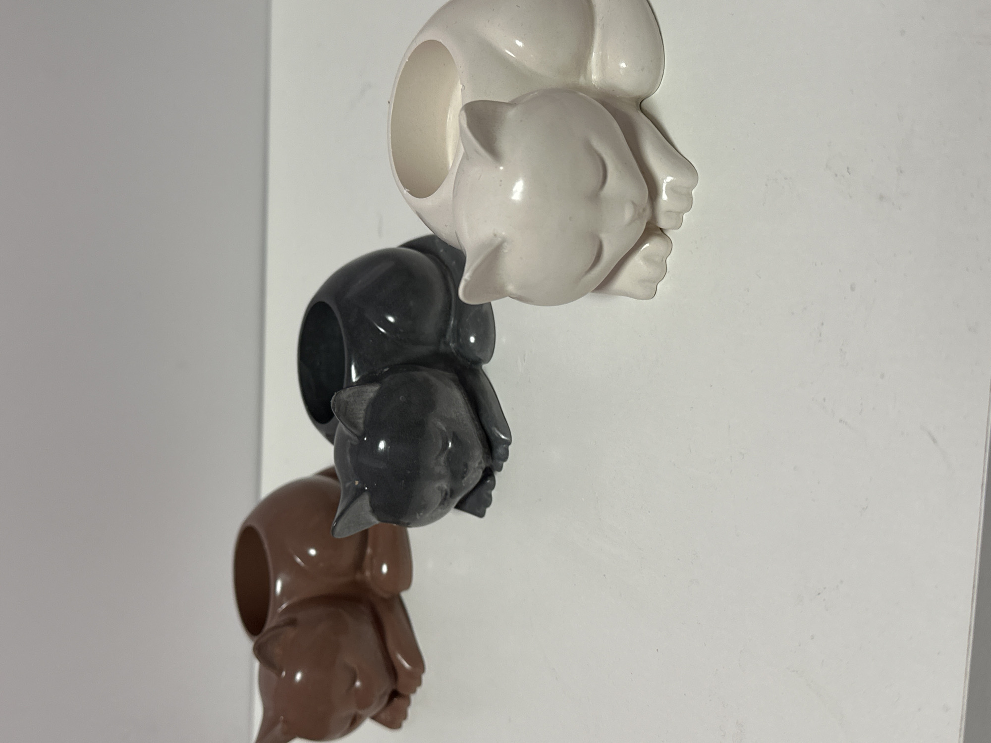 Three ceramic cat-shaped planters are arranged on a white surface. The planters are in different colors: brown, dark gray, and cream. They are all in a sleeping pose, with their eyes closed and paws tucked underneath them. The planters have an opening on their backs, suitable for holding small plants or other items. The background is a plain, light gray surface. The image is taken from a slightly elevated angle, looking down at the planters. There are no people or any other objects visible in the scene. The lighting is even, suggesting an indoor environment.