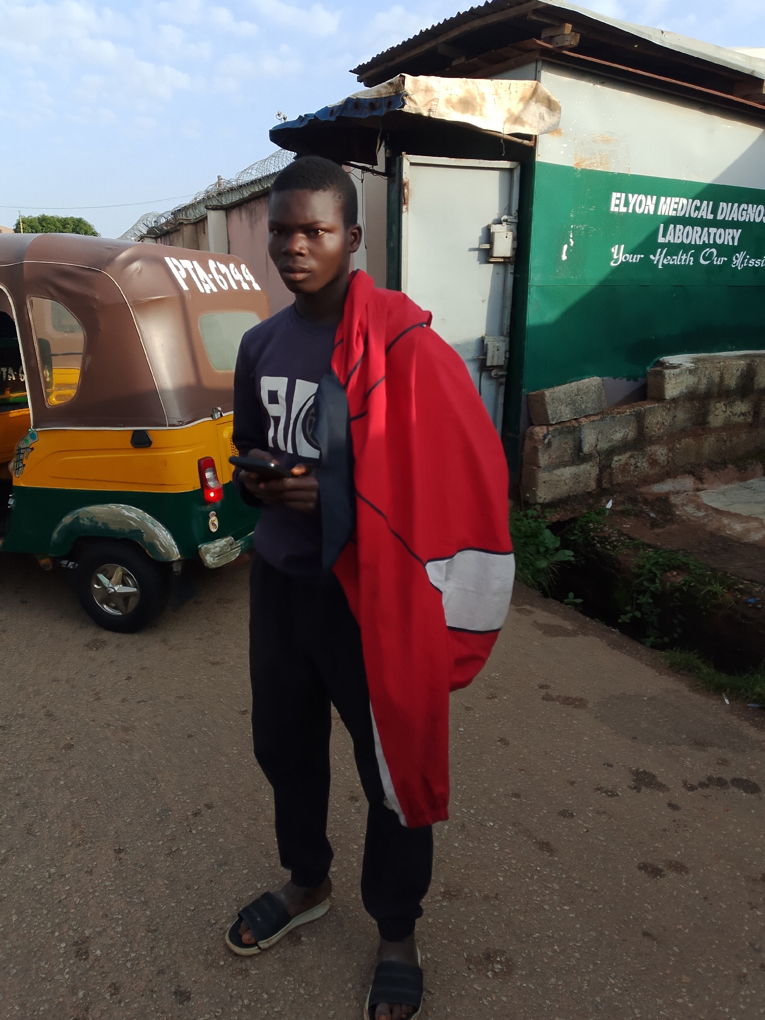 A young Black male stands on a paved street in front of a building with a green and white facade. The building has signage that reads "ELYON MEDICAL DIAGNOSTIC LABORATORY Your Health Our Mission". To the left of the male is a brown and yellow autorickshaw with "PTA 6744" visible on its side. The male is wearing a dark blue long-sleeved shirt with a white graphic on the front, black pants, and dark slide sandals. He is holding a black mobile phone in his right hand and has a red jacket draped over his left shoulder. The ground is asphalt and there are some weeds and a drainage ditch to the right of the building. The sky is overcast and appears to be early morning or late evening. The overall scene suggests a street-level view in a commercial or residential area of Jos, Nigeria.