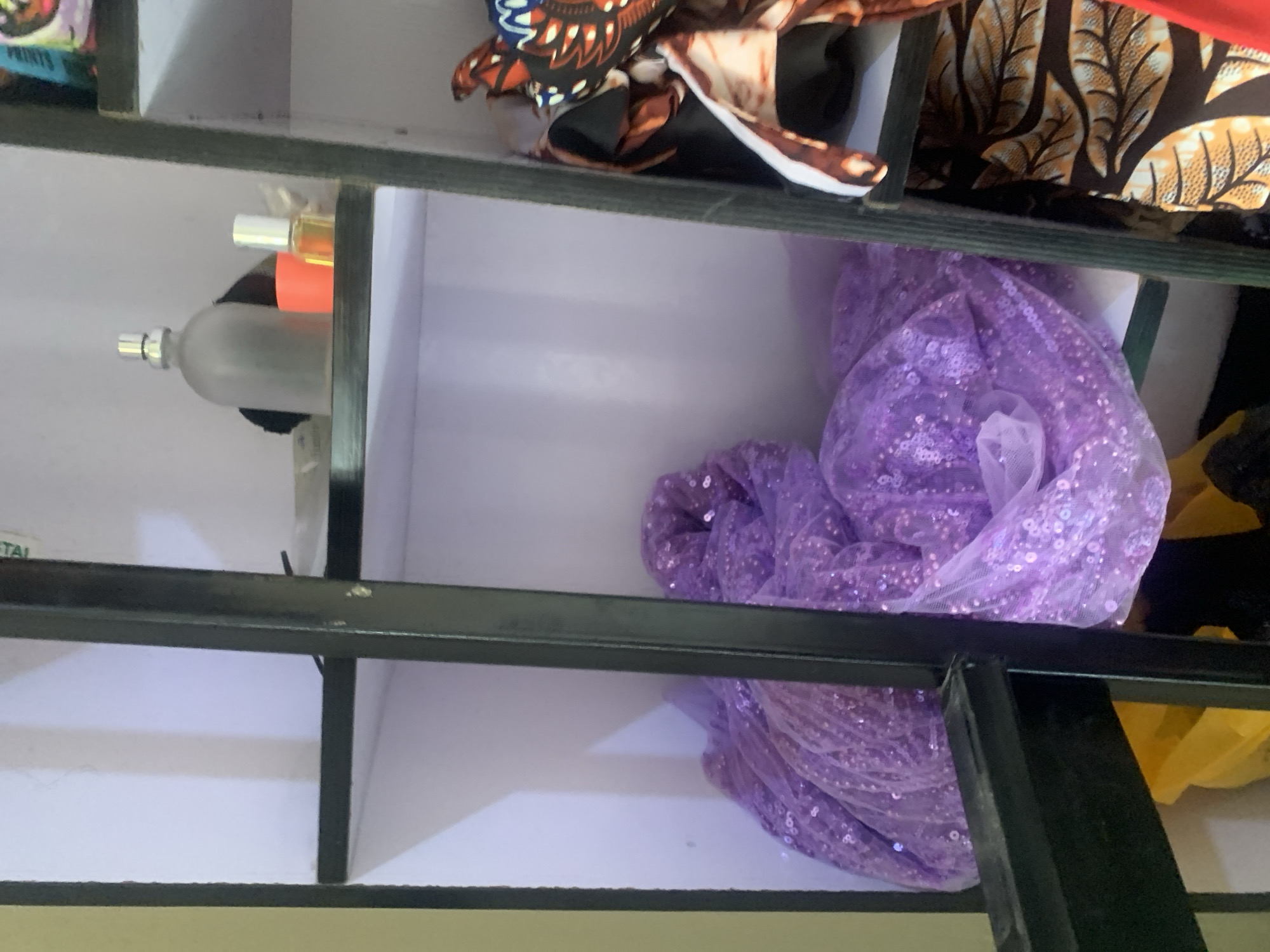 The image shows a shelf unit with several items stored on its shelves. On the top shelf, there are two bottles, one appears to be a perfume bottle, and a small orange container. Below this, a large piece of shimmering lavender fabric, possibly sequined tulle, is draped across the shelf. To the right of the lavender fabric, on a lower shelf, are several pieces of patterned fabric, including one with a bold brown and orange leaf print. The shelf unit is framed by black metal bars, and a glimpse of a yellow object, possibly a bag or container, is visible on the bottom shelf. The lighting suggests an indoor setting, likely a storage area or a retail space. The scene is located in Jauro Yinu, Nigeria.