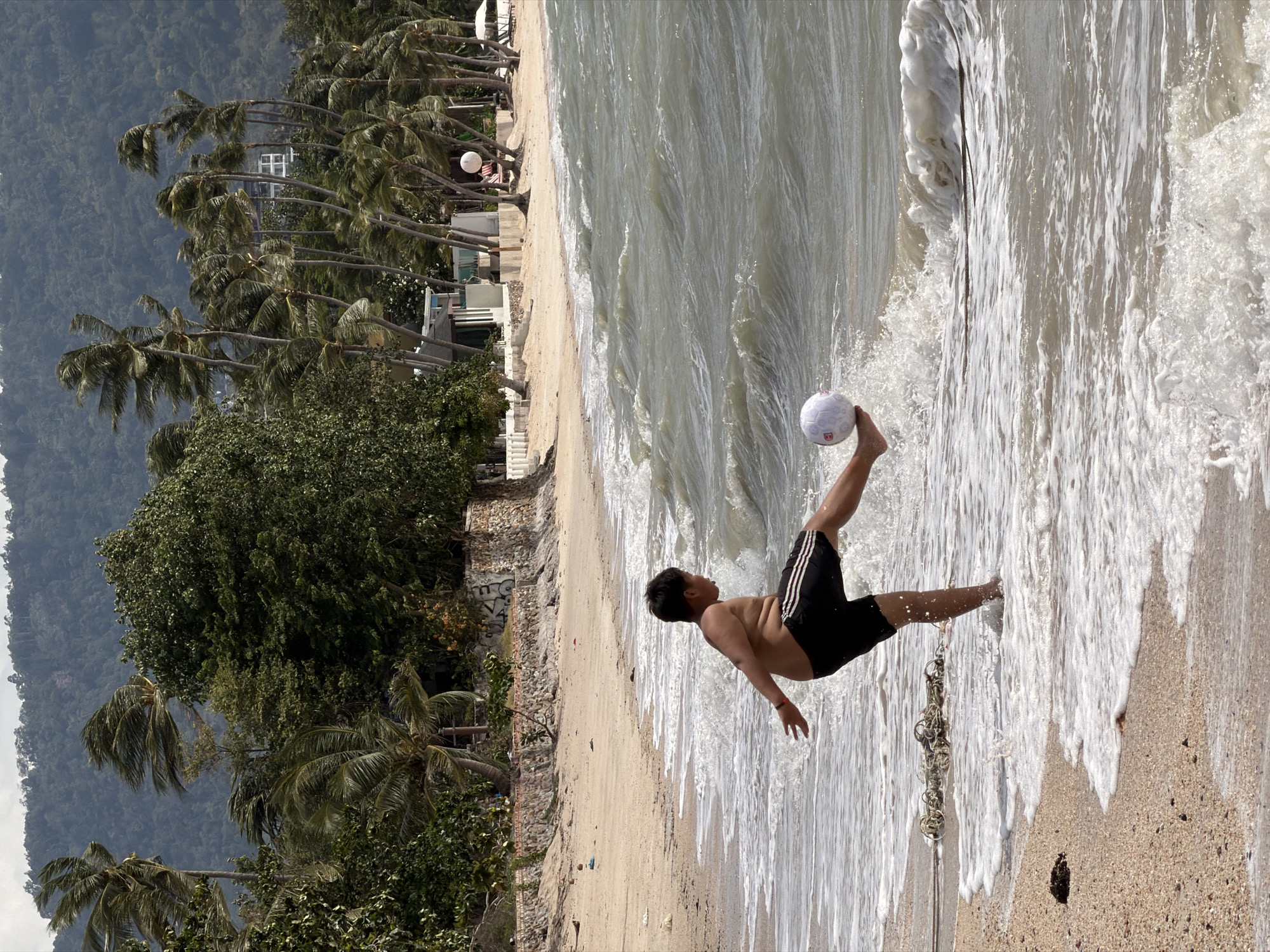 A young male is playing with a white soccer ball at the edge of the ocean on a beach, kicking the ball towards the water. He is wearing black shorts with white stripes. The beach has light-colored sand and is lined with palm trees and lush green vegetation in the background. A gentle wave is washing onto the shore, creating foam around the boy's feet. In the distance, buildings and a hilly, forested landscape are visible under a cloudy sky. The scene appears to be in the coastal region of Ko Samui District, Thailand, given the characteristic palm trees and tropical vegetation.