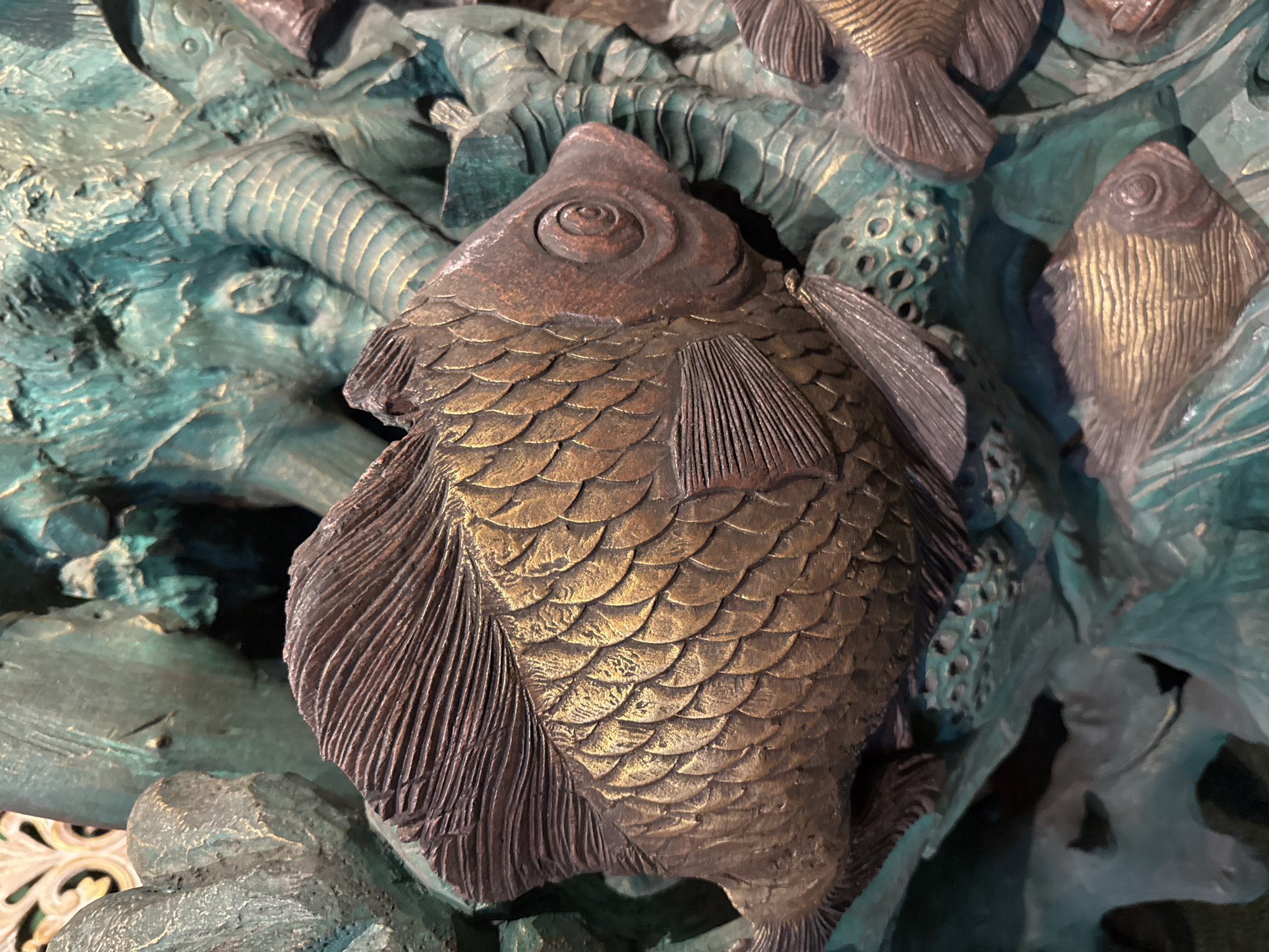 This is a close-up, angled shot of a bronze sculpture depicting fish amongst aquatic plants. The main subject is a large fish in the foreground, rendered in a mottled brown and gold patina, with intricately detailed scales and fins. To the right and bottom of the frame, smaller fish are visible, also with a similar bronze finish. The background consists of stylized, greenish-bronze representations of seaweed or coral, with a rough, textured surface. The lighting highlights the metallic sheen of the sculpture. The sculpture is located in Kuta, Indonesia.