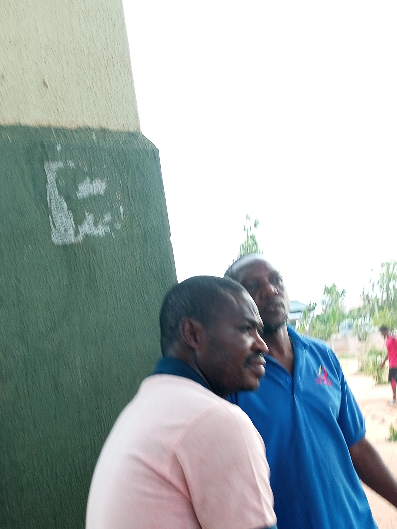 Two men stand near a green and cream colored wall. The man in the foreground is wearing a light pink polo shirt and the man behind him is wearing a blue polo shirt with a logo. The wall has a faint, smudged white marking that resembles the letter "E" on its green section. In the background, trees and buildings are visible, suggesting an outdoor urban environment. The sky is overcast.