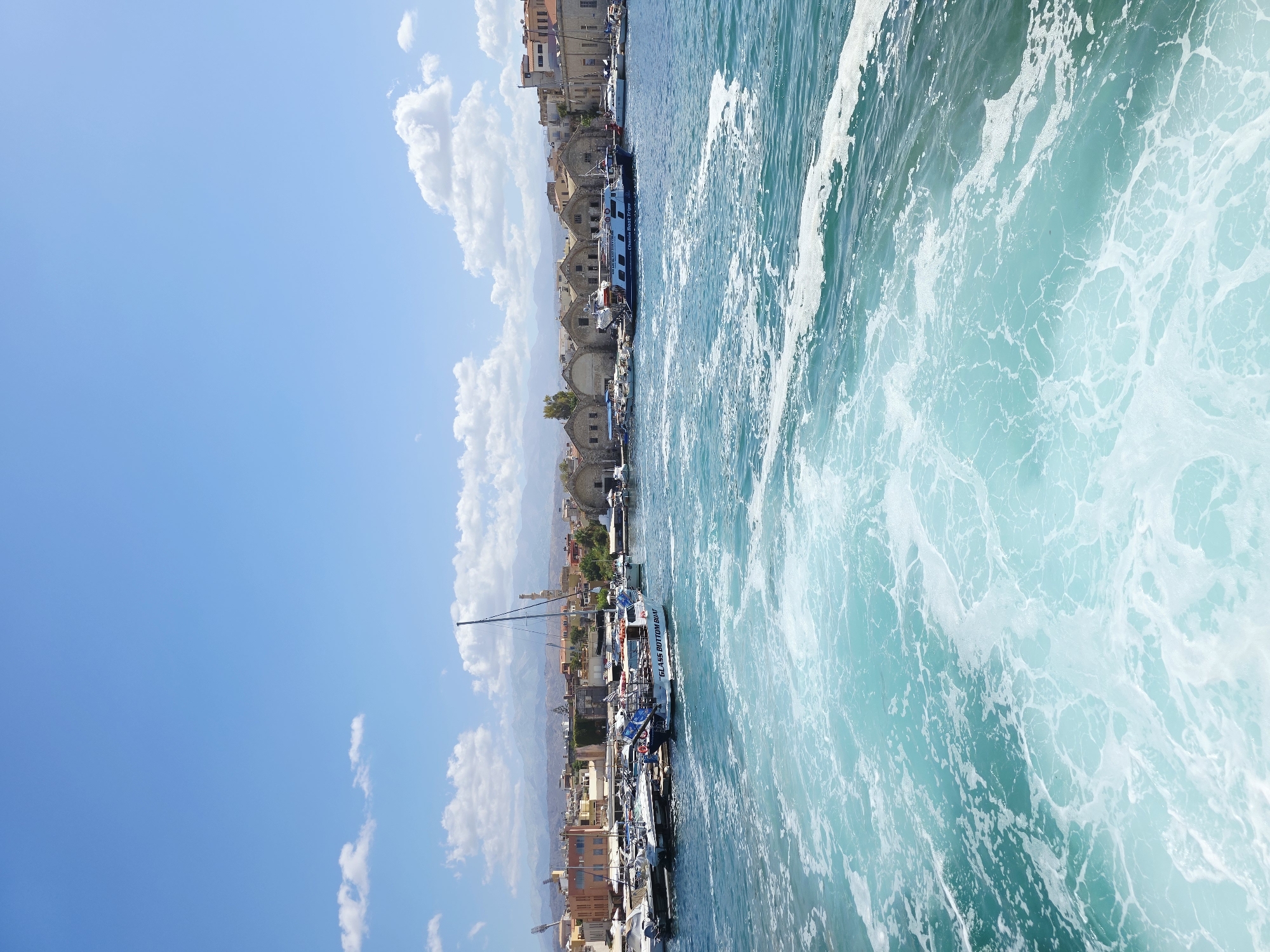 The image depicts a view of the Venetian Harbor in Chania, Greece, from the water. In the foreground, a trail of white foam churns in the turquoise water, indicating movement, likely from a boat. The middle ground features a collection of boats docked in the harbor. One prominent boat, a glass-bottom boat, has "GLASS BOTTOM BOAT" and "Chania Boat Trips" written on its side. Other smaller boats and a sailboat are also visible. The background showcases the historic waterfront buildings of Chania, with their characteristic architecture, and beyond them, hills and mountains under a bright blue sky dotted with white clouds. The scene is bathed in sunlight, suggesting a clear, pleasant day.