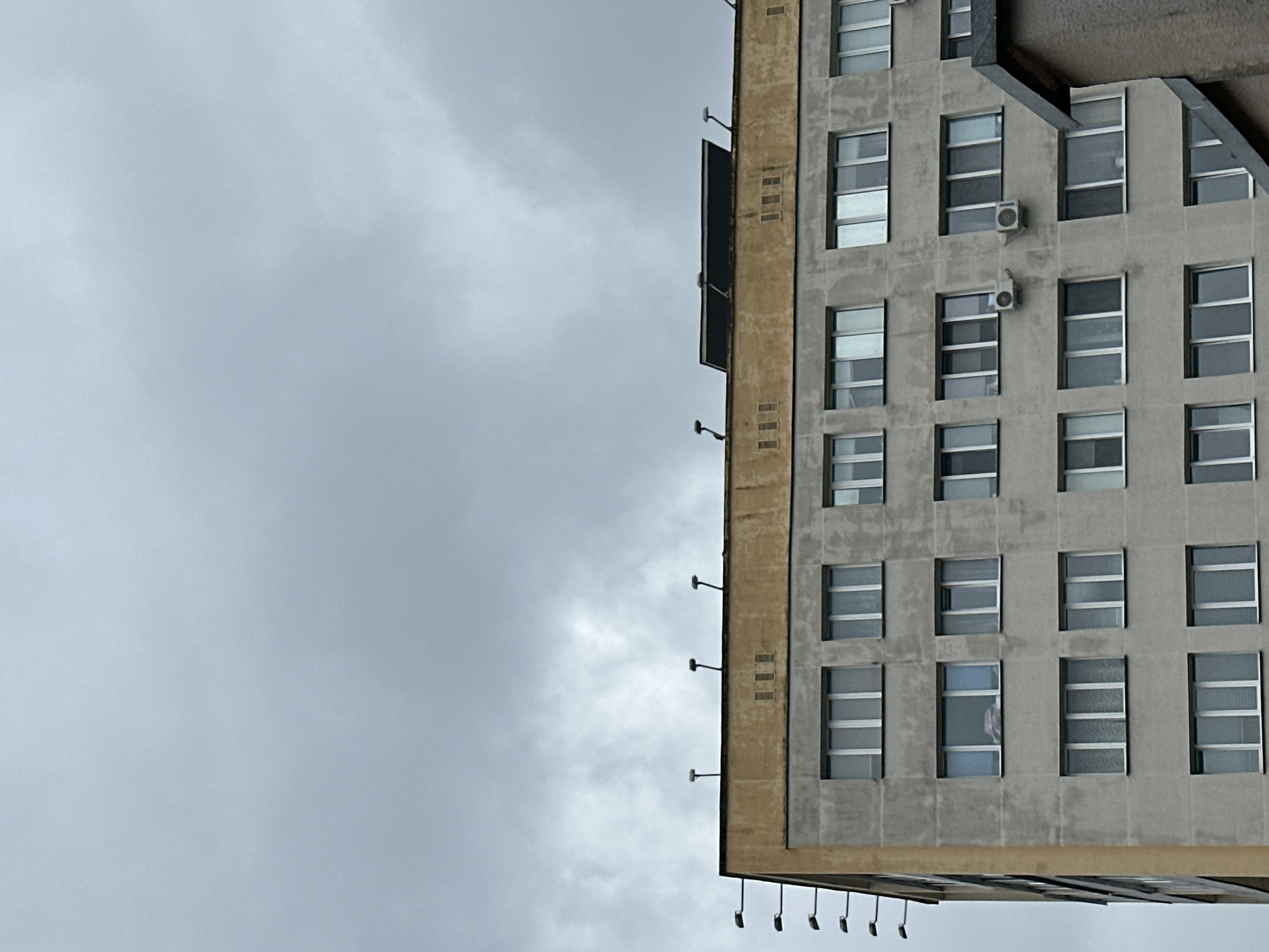 An exterior, eye-level, medium shot captures the upper portion of a monolithic concrete apartment building against a dark, overcast sky. The building, characterized by its repeating pattern of rectangular windows, is situated in Minsk, Belarus. The facade is a muted gray, showing signs of age and weathering, with a distinct yellow trim along the uppermost section. Several windows are visible on each of the building's visible floors, arranged in a grid. On the roofline, multiple streetlights are mounted on thin poles, and a dark, flat rectangular object, possibly a solar panel or a skylight, is also present. The sky is filled with dense, gray clouds, indicating a gloomy, possibly rainy day. A small portion of another building, with a dark, angular roof, juts into the frame from the right side. There are no people or visible signs of activity.