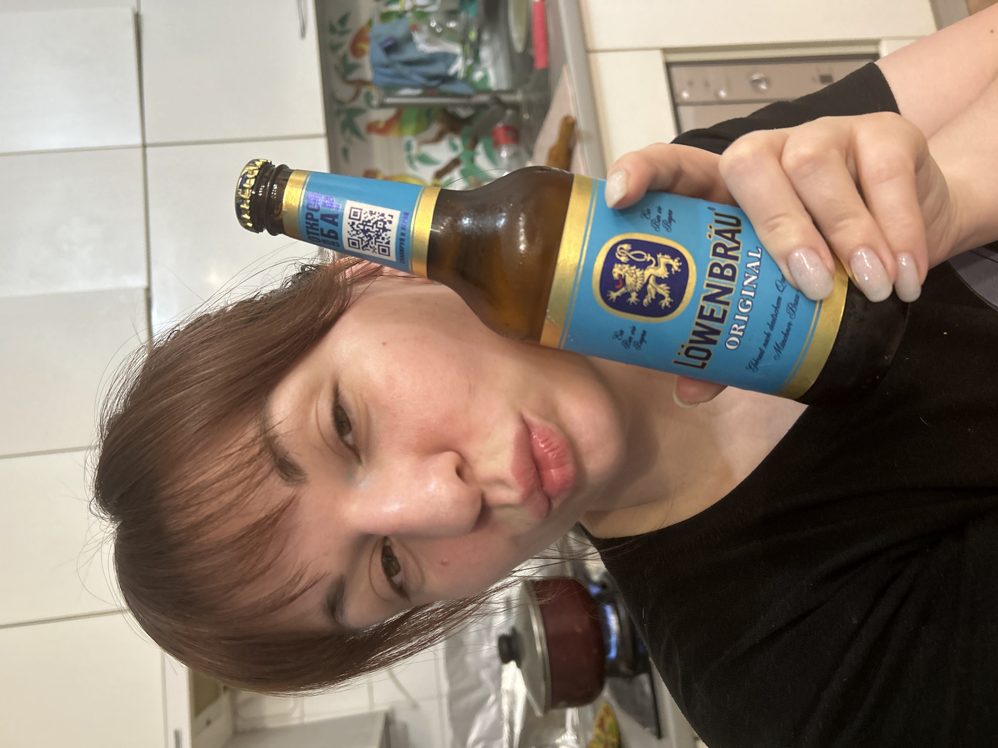 A woman is holding a bottle of Löwenbräu Original beer in front of her face. She has brown hair with bangs and is wearing a black shirt. The beer bottle has a blue label with a gold lion emblem. The label includes text in German, indicating it is brewed according to German quality standards. The background shows a kitchen setting with white tiles and various kitchen appliances. The label contains Russian text, suggesting it is sold in Фоки, Russia.