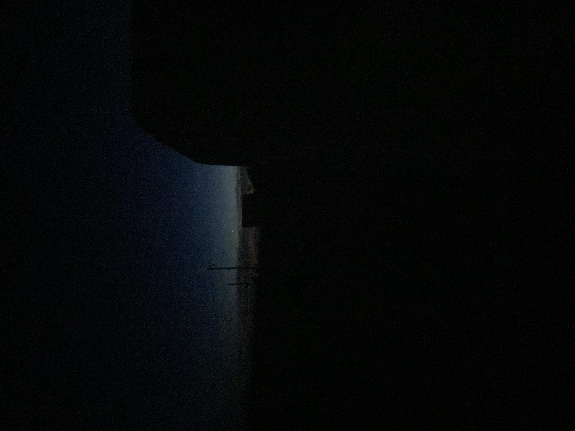 The image depicts a dark, grainy scene, possibly taken at night in the city of Прикамский, Russia. A body of water, possibly a river or bay, is visible on the left side of the frame with a faint, distant shoreline visible. There are a couple of structures on the coastline with what appear to be power lines leading to them. The right side of the frame is dominated by an area of complete darkness, perhaps a building or a hill. The top left also has some dark shapes, possibly a tree or a building roof.