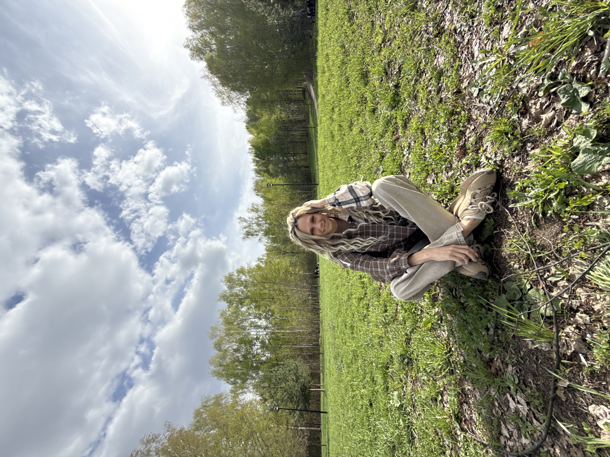 A woman is sitting cross-legged on a grassy area in a park, possibly in Rusanovo, Russia. She is wearing a plaid shirt, light-colored pants, and tan shoes. The background features trees and a cloudy sky.