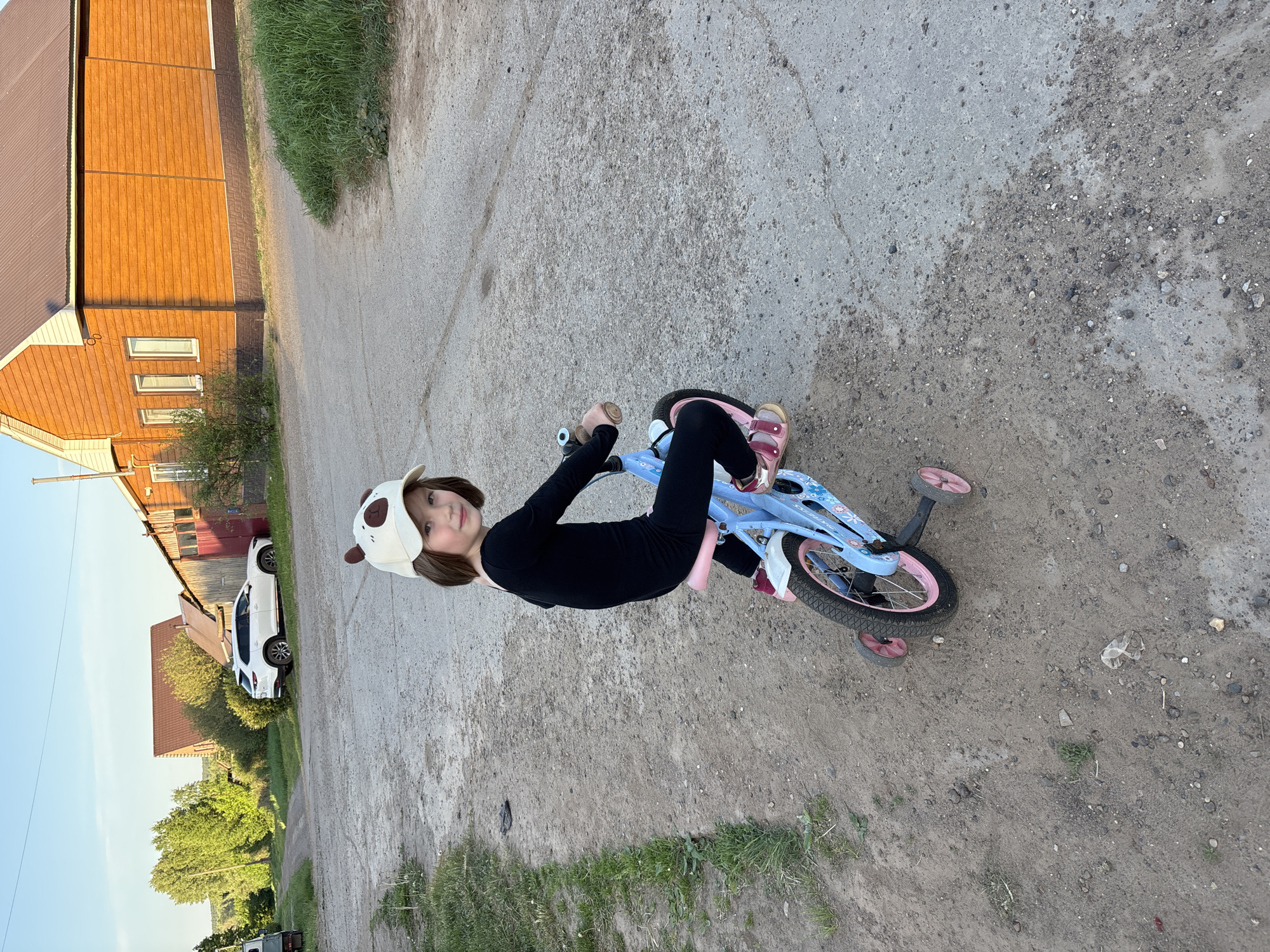 A young girl is riding a blue and pink bicycle with training wheels on a dirt road. She is wearing a black long-sleeved shirt, black pants, and pink sandals. The girl is also wearing a white and brown hat with animal ears. The background includes a tan and brown building with windows, a white car, green trees, and grass. The scene is in Niva, Russia.