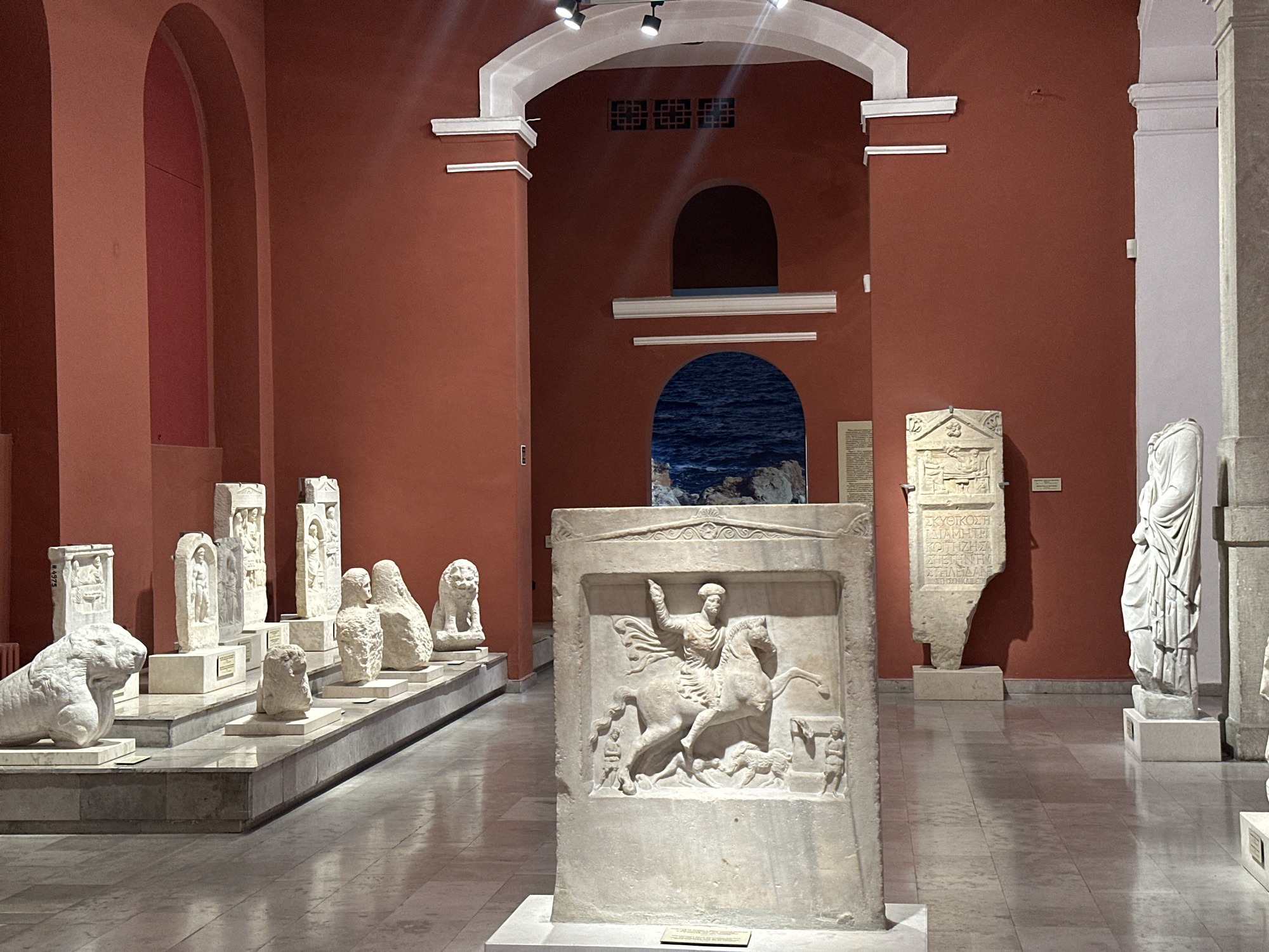 This image depicts an exhibit within the Varna Archaeological Museum in Varna, Bulgaria. The scene is an indoor museum hall with deep red walls and high ceilings, featuring arched doorways and architectural elements suggestive of classical design. The floor is tiled in a light gray and white checkered pattern, reflecting the overhead lighting.
Dominating the foreground is a large, intricately carved stone stele that stands upright on a light-colored pedestal. The stele features a relief sculpture of a man on horseback, with other figures and animals below. To the left of this central piece, a row of ancient stone artifacts is displayed on tiered pedestals. These artifacts include several upright stelae with reliefs and inscriptions, as well as smaller sculptures, including a lion and what appears to be a human bust.
Further to the right, another upright stone slab with an inscription is visible. Adjacent to it stands a partial marble statue of a robed human figure, and behind it, another indistinct stone sculpture. In the background, through an archway, a depiction of the sea with rocks can be seen, possibly a mural or a framed artwork. The lighting in the museum is a mix of spotlights from above and ambient light, casting shadows on the floor and walls. The overall impression is one of a well-curated archaeological exhibition.