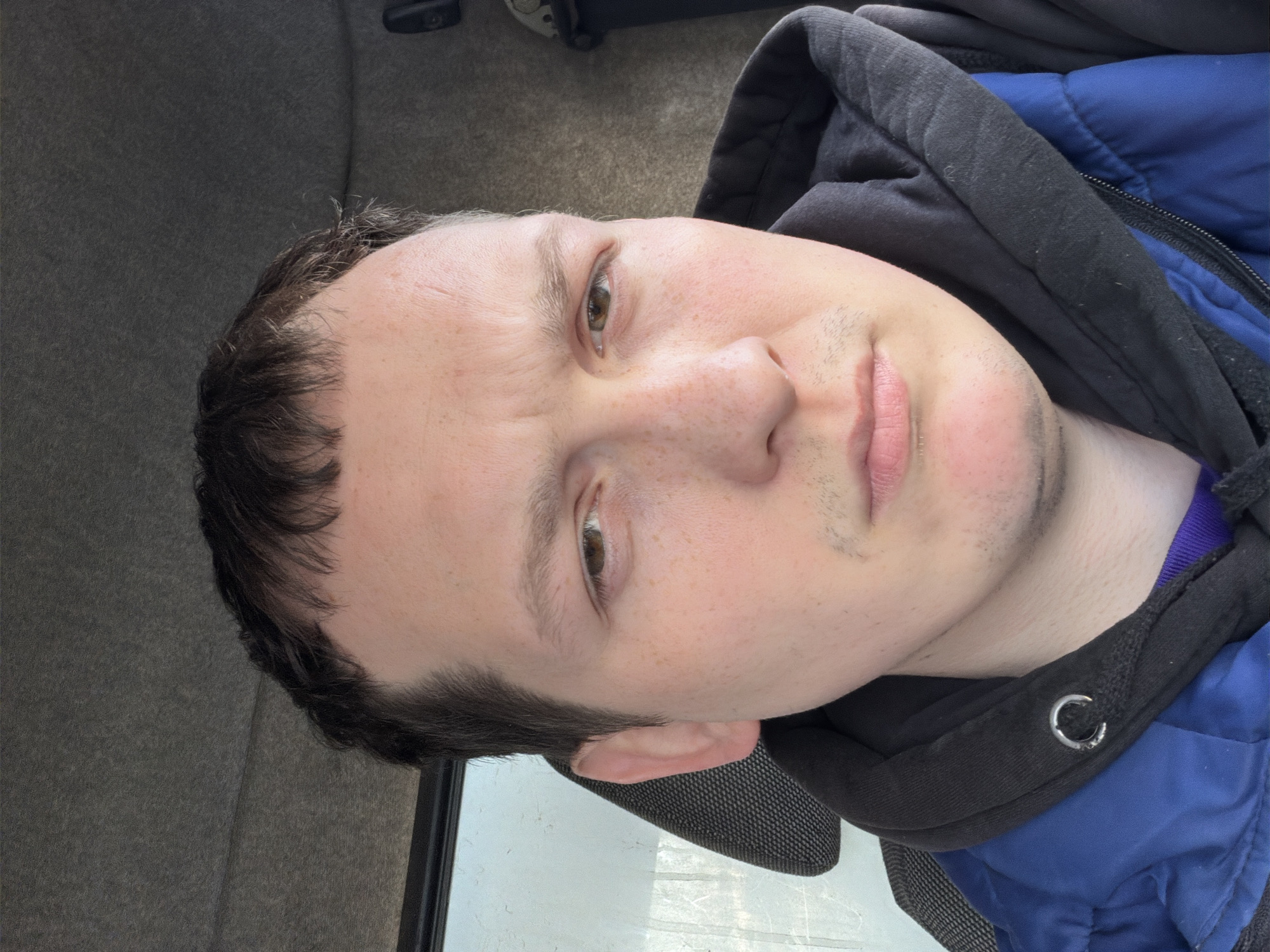 A close-up, slightly angled portrait of a fair-skinned person with short, dark brown hair and hazel eyes. The person has a few freckles on their face and slight stubble. They are wearing a blue jacket with a black hooded sweatshirt underneath, the hood visible around their neck. The background consists of grey fabric, suggesting an interior, possibly a car. There is no information to suggest the location is in the city of Chaikovsky, Russia.