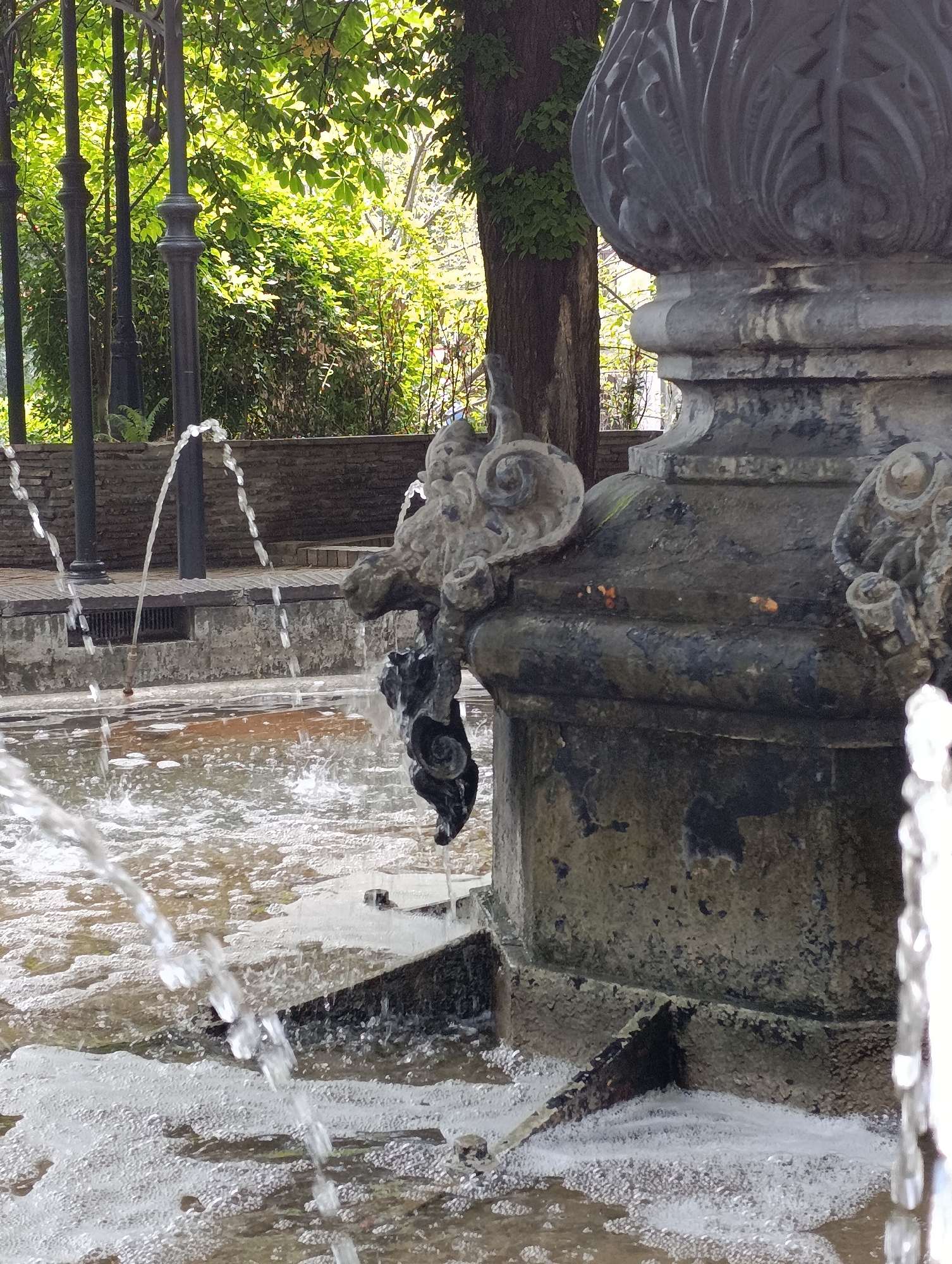 The image shows a close-up view of a fountain in Kyiv, Ukraine. The fountain is made of stone and has decorative carvings. Water is spraying from the fountain into a pool below. The pool is filled with water and foam. In the background, there are trees and a brick wall. The lighting suggests it is daytime.