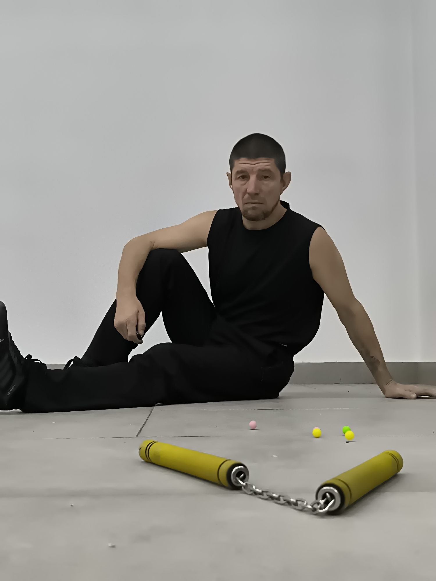 A man in a black sleeveless shirt and black pants sits on a tiled floor in front of a plain white wall. His left leg is bent with his foot tucked towards his body, and his right leg is extended out. His left arm is bent at the elbow, resting on his bent knee, while his right arm is extended back, hand resting on the floor. He has short, dark hair and a beard and is looking directly at the camera with a neutral expression. In the foreground, a pair of yellow nunchucks lies on the floor, connected by a chain. Nearby, several small, colorful balls are scattered on the floor. The environment appears to be an indoor space, possibly a training facility or an empty room, given the lack of other objects or decorations. The lighting is even and casts no harsh shadows.