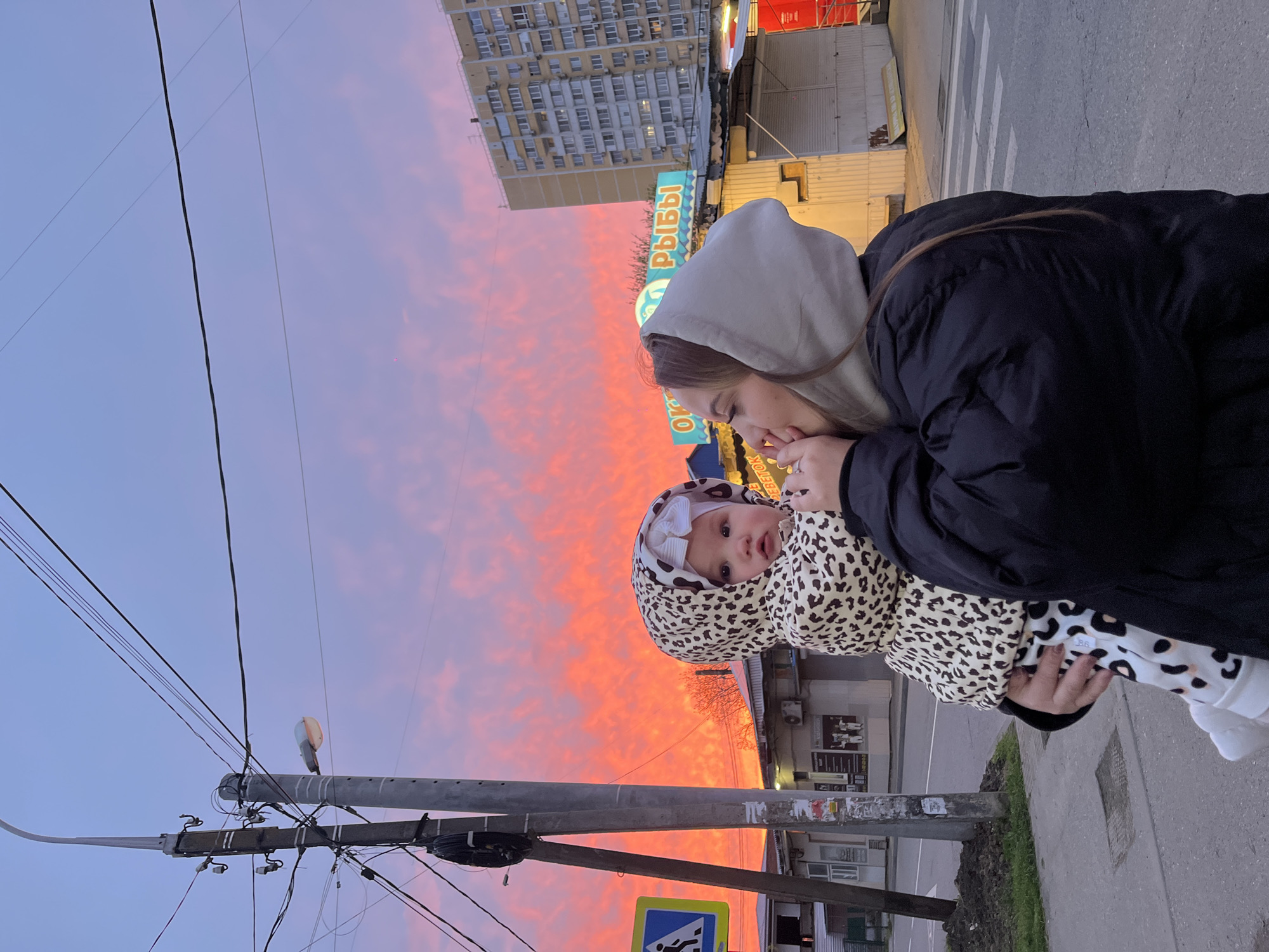 This image, set in Krasnodar, Russia, captures a tender moment between a woman and a baby outdoors during sunset. The sky dominates the upper portion of the frame, ablaze with vibrant pink and orange clouds that transition into a soft blue, indicative of either dawn or dusk.
A woman, dressed in a black puffer jacket and a light grey hooded sweatshirt, holds a baby or toddler. She looks down gently towards the child. The baby, wearing a distinctive leopard-print hooded outfit with a white bow on its head, is looking towards the camera with a calm and curious expression.
The setting is an urban street scene. In the foreground on the left, a utility pole laden with multiple power lines stands prominently. Below it, a blue pedestrian crossing sign is visible. The ground shows a paved sidewalk next to a street with white crosswalk markings. In the background, various buildings line the street. A tall, multi-story apartment complex is seen on the left, while commercial structures, one featuring a teal-colored sign with the Russian word "РЫБЫ" (RYBY), meaning "FISH," are present on the right. The scene is imbued with a tranquil atmosphere, contrasting the intimate human subjects with the expansive, colorful sky and typical city environment.