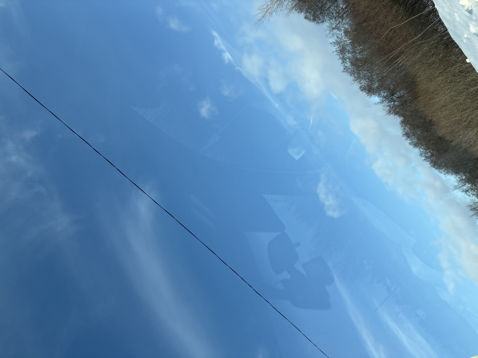 A reflective scene reveals a blue sky with scattered clouds, possibly viewed through a car window due to the reflection of interior features. A thin power line cuts diagonally across the left side. A dense treeline with snow can be seen on the right, suggesting a winter landscape. The composition is slightly tilted.