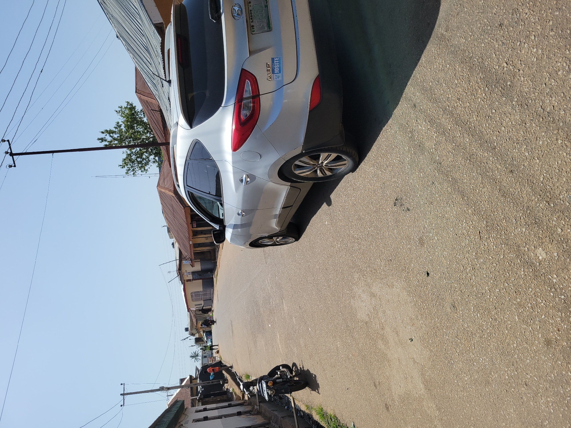 A silver Hyundai SUV is parked on the side of a street, with a motorcycle parked on the opposite side. The street is paved and lined with buildings, some with corrugated iron roofs. Power lines stretch above the street. The location is the city of Unknown, Unknown.