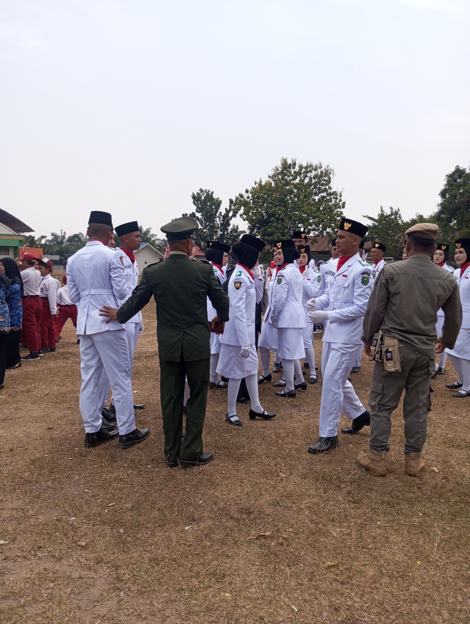In Pematang Reba, Indonesia, a group of people, primarily young individuals in white uniforms with red sashes and black hats, stand on a dry, grassy field, likely during an outdoor event. The uniforms suggest a formal gathering, possibly a school assembly or a ceremonial occasion. One person, in a dark green military-style uniform, faces away from the camera, appearing to interact with a young person in a white uniform. Another individual, in a khaki uniform and hat, stands to the right, also facing the group. In the background, several buildings and trees are visible, hinting at an open area within a community. The sky is overcast, indicating a cloudy day. The overall scene depicts an organized gathering with elements of uniformed personnel and participants, set against a backdrop of typical Indonesian outdoor scenery.