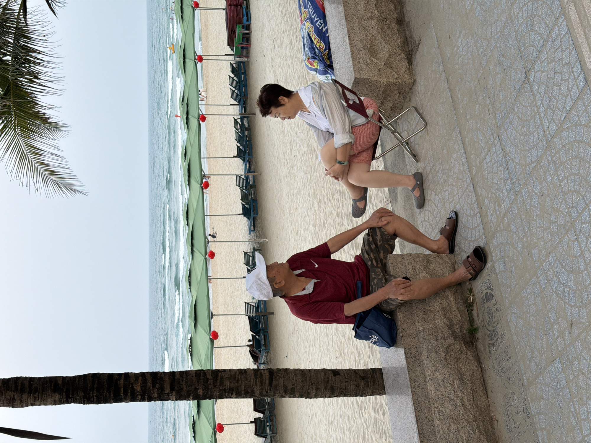 This image depicts a beach scene in Đà Nẵng, Vietnam. In the background, there is a view of the beach, ocean, and sky. Rows of green beach umbrellas with red accents line the sand. In the foreground, a woman sits on a folding chair, and a man sits on a stone ledge. Both are facing the beach and appear to be relaxing or observing the scene. The image also includes a palm tree trunk on the left side and tiled ground on the right side.