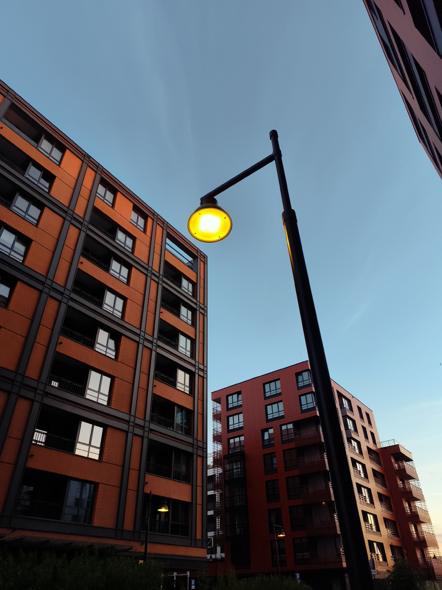 The image shows a modern residential building with an orange brick facade and dark gray structural elements against a clear blue sky. The building features multiple floors with numerous windows and balconies. A black lamppost with a bright yellow light is positioned in the foreground, angled upwards. Another building, also with an orange facade, is visible in the background to the right. The perspective is from a low angle, looking up at the structures. Based on the architectural style and common urban landscaping in Poland, this scene is likely located in the city of Gdańsk, Poland.