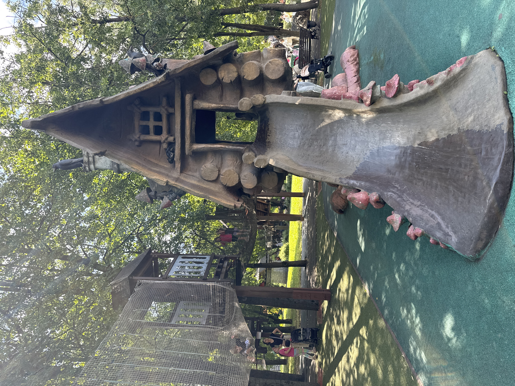 The image depicts a whimsical playground designed to resemble a fairytale village, set against a backdrop of lush green trees and a bright sky, suggesting a daytime scene. The focal point is a large, fantastical house-like structure with a steeply pitched roof and log-like siding, from which a wide, grey slide descends. The slide is flanked by decorative elements resembling oversized, pink mushrooms. To the left of this main structure, a multi-level play area is visible, connected by netted walkways and bridges, with children actively engaged in playing. Various adult figures are also present, some supervising, others pushing strollers. The ground surface of the play area appears to be a soft, green mat, interspersed with patches of grass and sunlight dappled by the overhead foliage. The overall impression is one of a vibrant and engaging children's recreation space. This playground is located in the city of Sovkhoz imeni Lenina, Russia, a settlement known for its well-maintained public spaces and unique architectural features, including this fairytale-themed attraction.