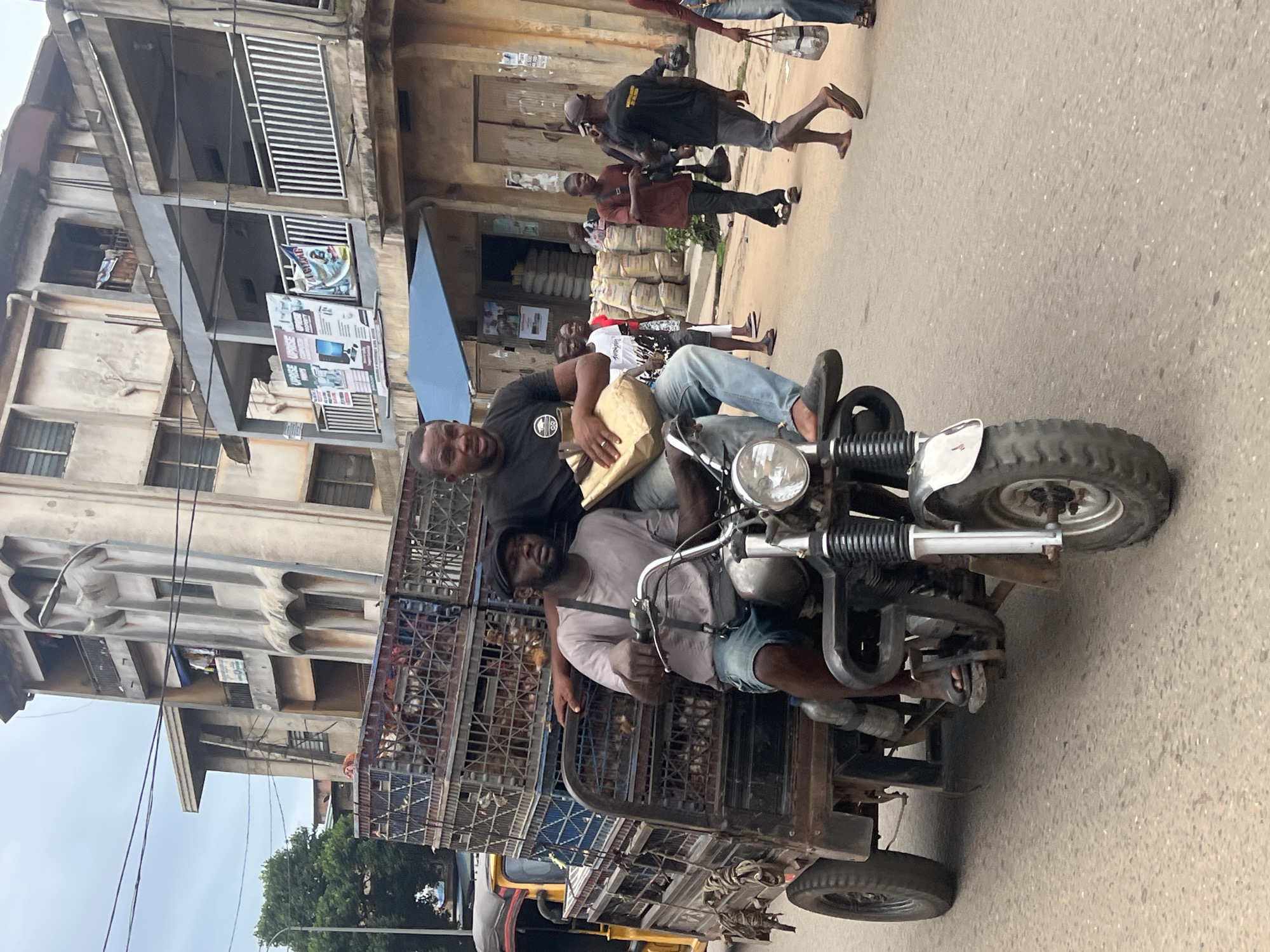 In Nkpor, Nigeria, two men are riding on a three-wheeled motorcycle loaded with cages of poultry. The man in the front is driving, wearing a grey t-shirt and denim shorts, with a black bag slung across his chest. The man behind him, wearing a black t-shirt and jeans, is holding a light brown bag. The motorcycle's cargo area is stacked with multiple large, metal cages, each containing numerous chickens. In the background, a multi-story building with balconies and windows lines the street. Several other people are visible walking on the pavement and in the street, some carrying bags. The scene appears to be a bustling urban environment.