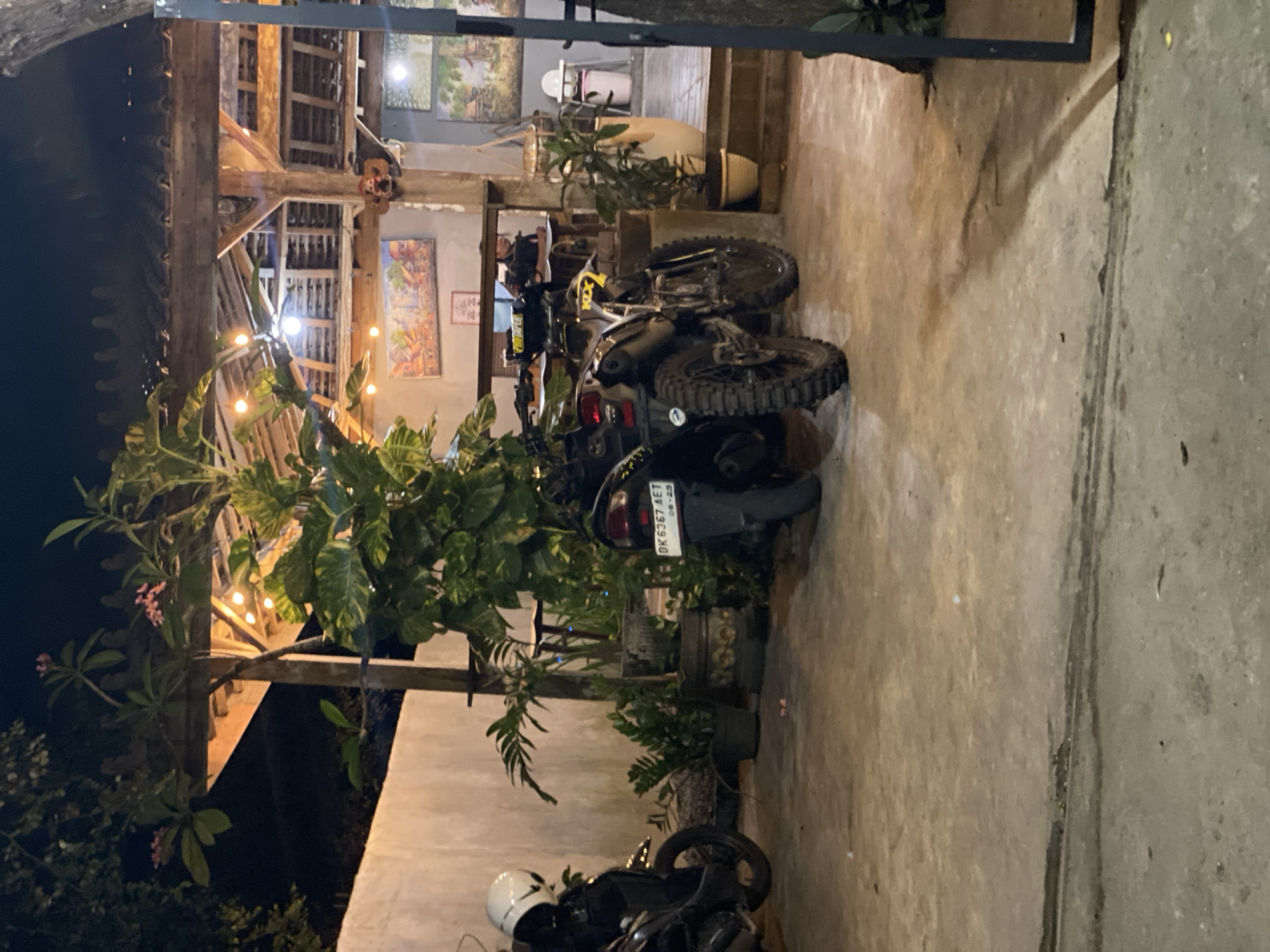 The image captures an outdoor scene at night, likely a restaurant or cafe in Kuta Selatan, Indonesia, illuminated by string lights under a wooden pergola. In the foreground, two motorcycles are parked on a concrete surface. The motorcycle on the left is mostly obscured, with only its front wheel, a white helmet on its seat, and part of its frame visible. The motorcycle on the right is a dirt bike with knobby tires, facing towards the right side of the frame. It has a yellow and black color scheme and a license plate reading "DK 6367 AET".
Behind the motorcycles, a lush green plant with large leaves dominates the center of the frame, with smaller plants in pots scattered around. In the background, a wall is visible on the left, and a building with a wooden structure and artwork on the walls is seen further back. A person can be seen partially, sitting behind a counter in the background. The overall atmosphere is relaxed and tropical.