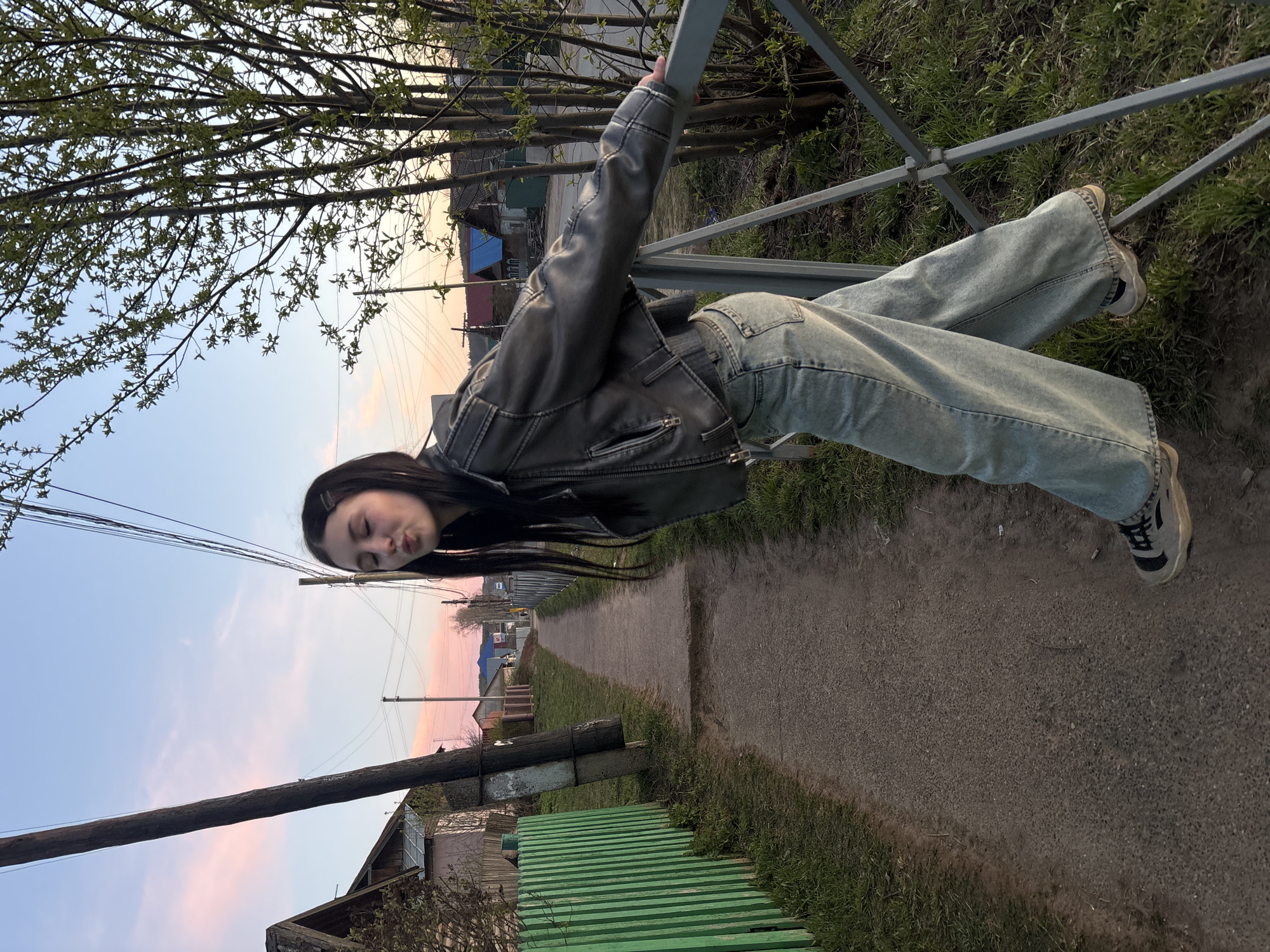 The photograph shows a young woman hanging from a metal railing along a paved road in Фоки, Russia. She is wearing a dark leather jacket, light-wash denim pants, and sneakers. She has long, dark hair. The road runs between green grass and a few houses. Above her is a sky with pale pink and blue tones. A wooden utility pole is visible on the left side of the image. Bare branches of trees are at the top of the frame.
