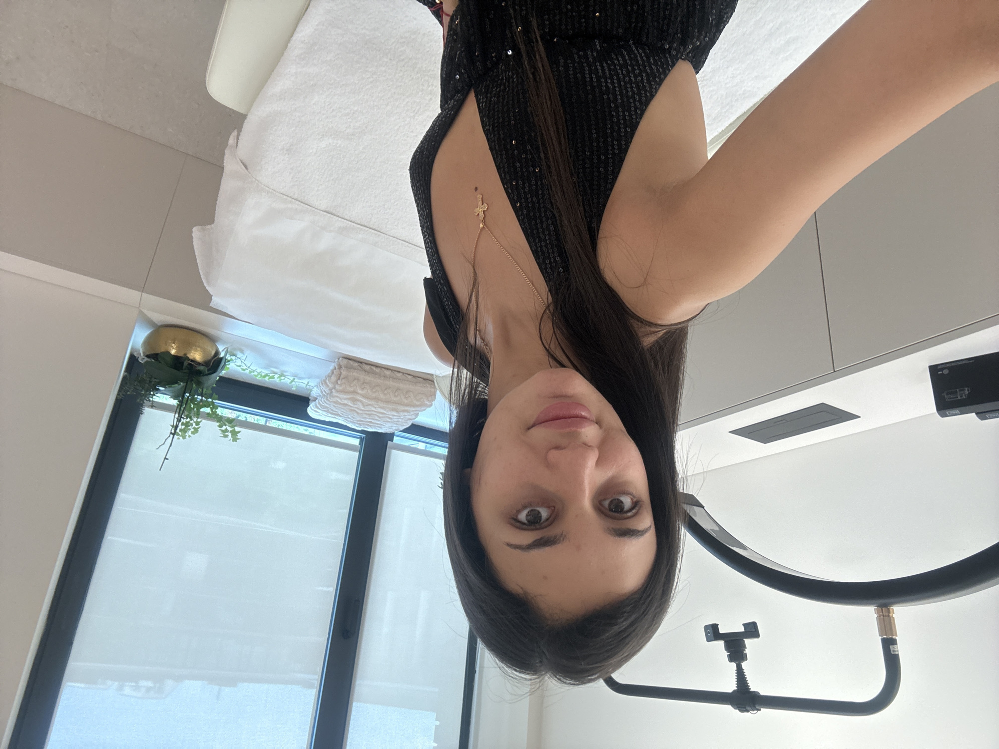 A young woman with long dark hair, wearing a sparkling black dress with a deep V-neck and a gold cross necklace, is taking a selfie in a brightly lit, modern room. She is positioned in the foreground, with her right arm extending towards the camera. Behind her, to the left, is a white counter with a black device. To her right, there is a white massage table covered with a white sheet and towels. Further back, a large window with blinds is visible, and on the windowsill, a small potted plant sits in a golden pot. The overall impression is that of a clean, minimalist interior, possibly a spa or a treatment room. There is no indication that this location is in Sofia, Bulgaria.