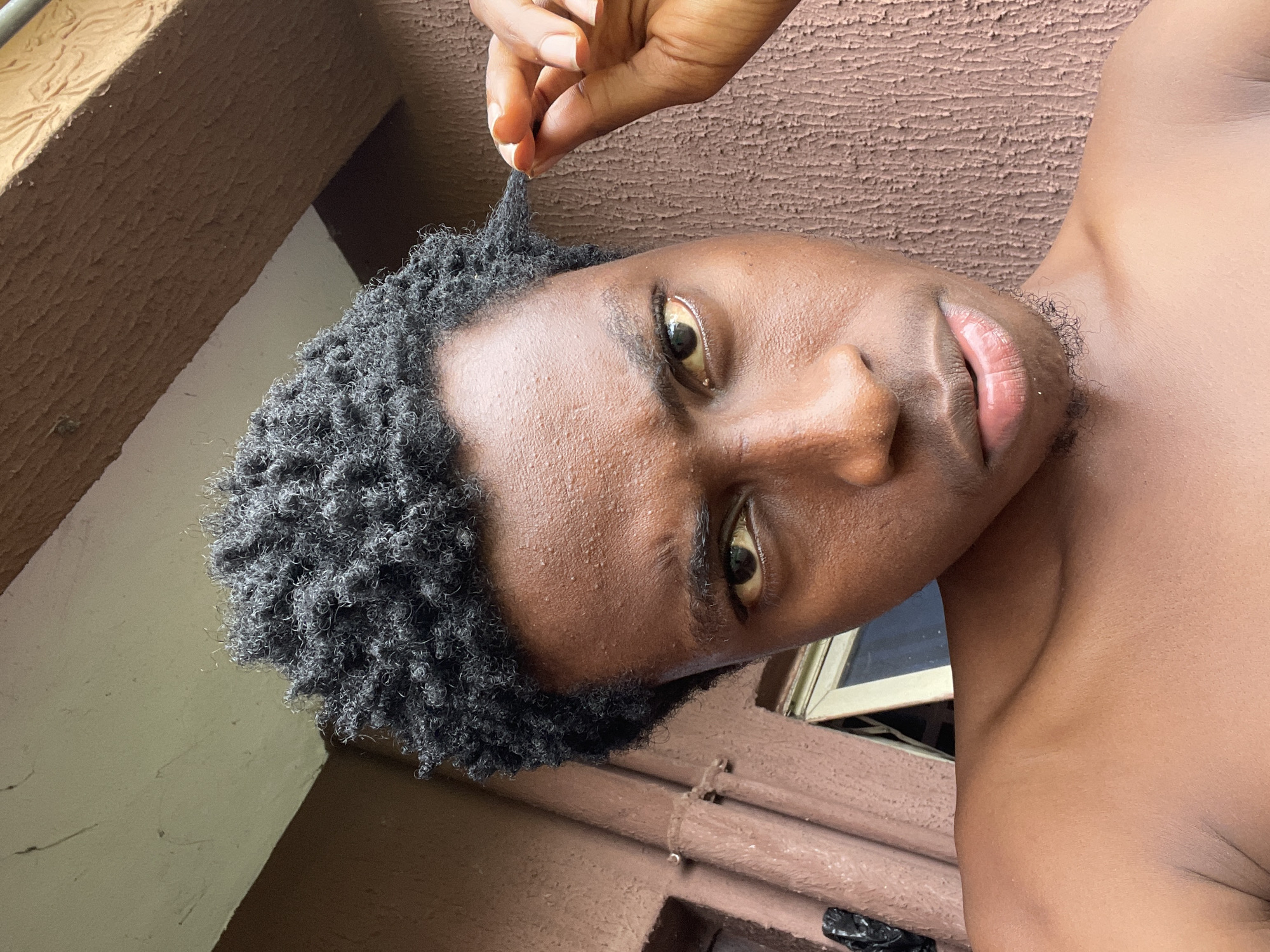 A medium shot captures a young Black man from the chest up, looking directly at the camera with a concerned expression. He has dark, curly hair and is wearing no shirt. His right hand is raised to his hair, gently pulling a lock. The background is a textured brown wall, possibly the exterior of a building. To the left, a window frame and part of a brown downspout are visible. The scene appears to be outdoors, with soft, natural light illuminating the man's face. The location is specified as Nkpor, Nigeria.