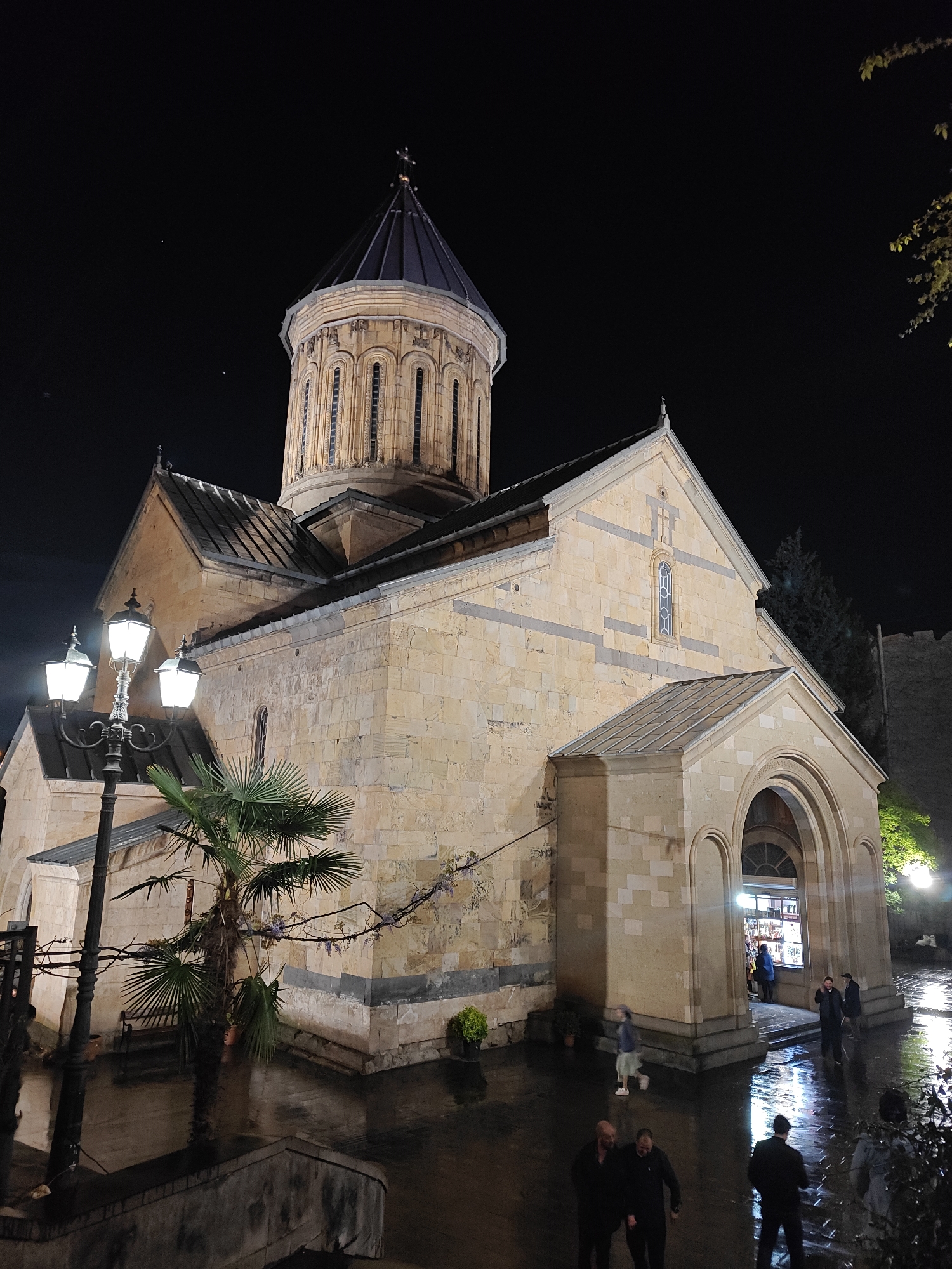 This night-time photograph from Tbilisi, Georgia, features a well-lit Georgian Orthodox church as its central subject. The church is constructed from light-colored stone blocks, characterized by a large, ornate dome with a conical roof and a cross at its apex. Its facades exhibit arched windows, some stained glass, and decorative stone banding.
In the foreground, an ornate black street lamp with three bright lanterns illuminates the scene. Directly beneath the lamp, a small palm tree stands beside the church's stone wall. The ground in front of the church is wet and reflective, suggesting recent rain. Several people are visible in the lower half of the image, some walking and others standing in small groups, their reflections shimmering on the wet pavement.
One of the church's arched entrances on the right side houses a brightly lit shop or kiosk, displaying various items, with a person visible inside. A darker, more rugged wall or structure is partially visible in the background to the right of the church, partially obscured by trees. Tree branches with some light-colored leaves extend into the upper right corner of the frame. The dark night sky provides a stark contrast to the illuminated church and street scene, emphasizing the artificial lighting. No clear text is legible in the image.