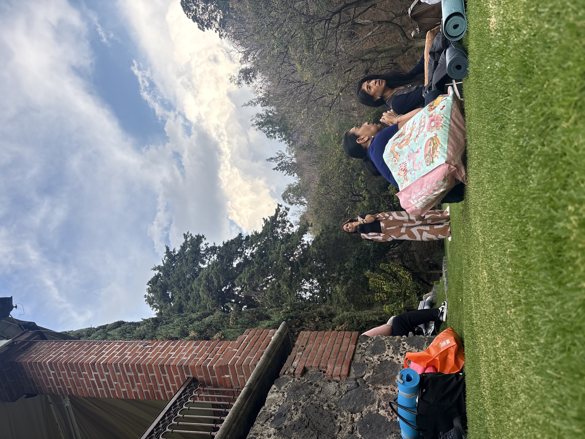 The scene shows three women resting on a grassy hill in Santa Ana Acozautla, Mexico. Two are lying on a blanket and yoga mats, looking up at the sky, while the third woman stands above them. A brick and stone wall is visible in the foreground, with trees and sky in the background. Personal items like bags and additional yoga mats are scattered around the base of the hill.