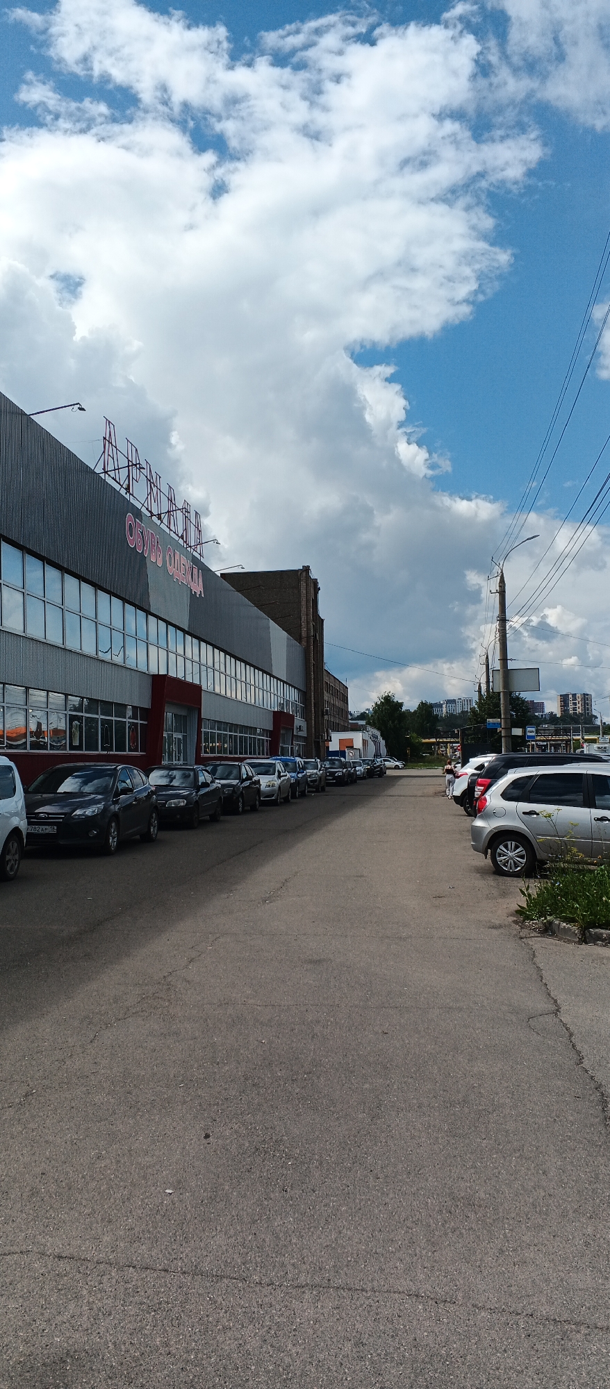 The image shows a street scene in Izhevsk, Russia, with a large building on the left that appears to be a retail store. The building has a metal facade and a large sign that reads "АРМАДА" in red letters at the top, and "ОБУВЬ ОДЕЖДА" (Shoes Clothing) below it. A row of cars is parked along the street in front of the building. The street is paved and there are trees and other buildings in the background. The sky is partly cloudy with a mix of blue sky and white clouds. The scene is captured from a low angle, looking towards the building and down the street. There are no people clearly visible in the image.