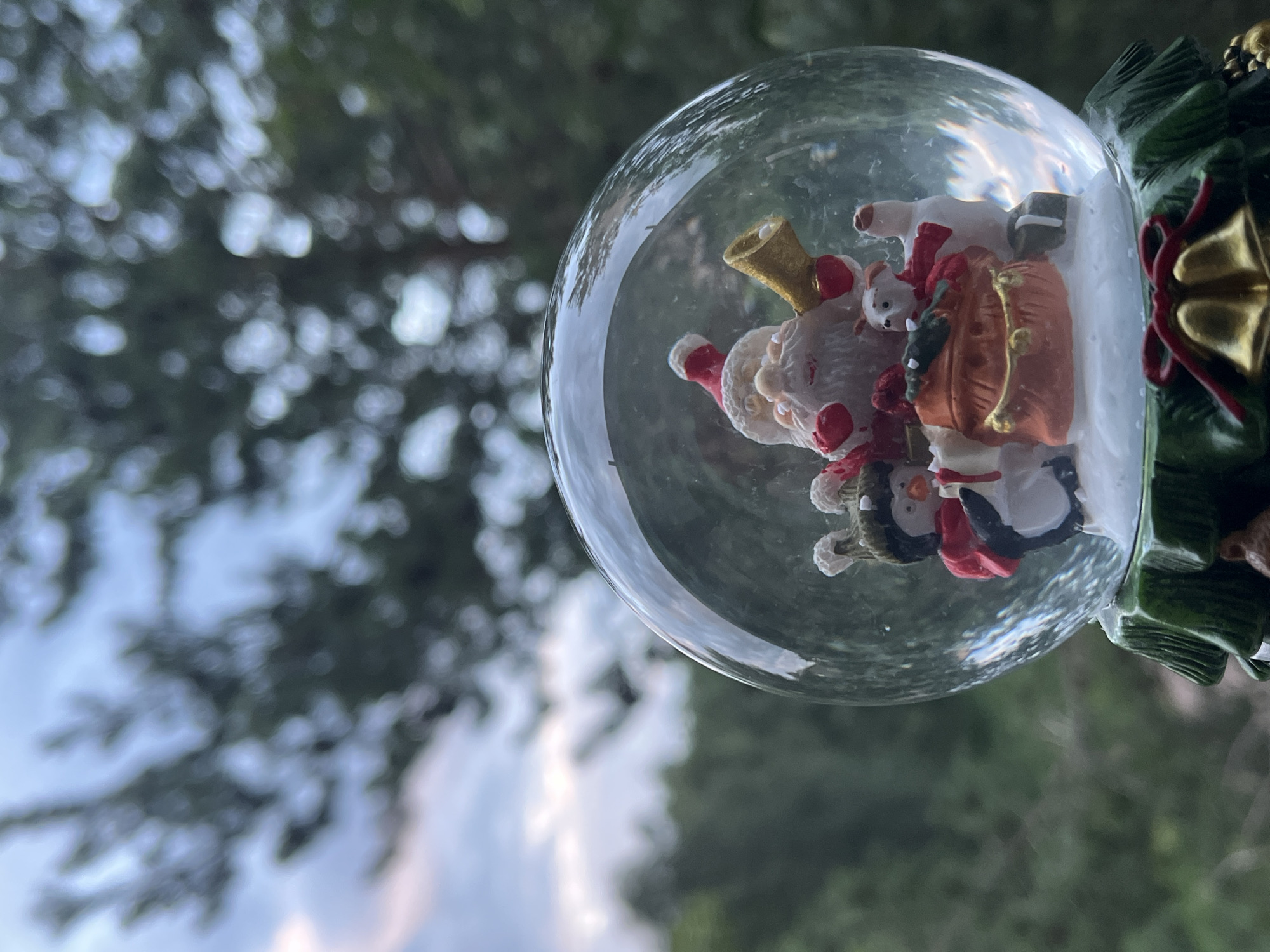 This media file is a close-up, eye-level shot of a Christmas snow globe. The snow globe features a figurine of Santa Claus, along with two penguins and a polar bear, all arranged in a festive scene. Santa is wearing his traditional red and white suit and hat, and is holding a sack. The penguins are also dressed in festive attire, with one wearing a green knitted hat and a red scarf, and the other with a red scarf. The polar bear is white with red accents. The base of the snow globe is decorated with green foliage and gold bells, adding to the Christmas theme. The snow globe is partially submerged in what appears to be water or glitter, obscuring some of the details. The background is blurred, suggesting an outdoor setting with green foliage and a cloudy sky, hinting at a possible daytime scene with overcast weather. The focus is sharp on the snow globe and its contents, while the background is soft and out of focus. There is no visible text in the image.