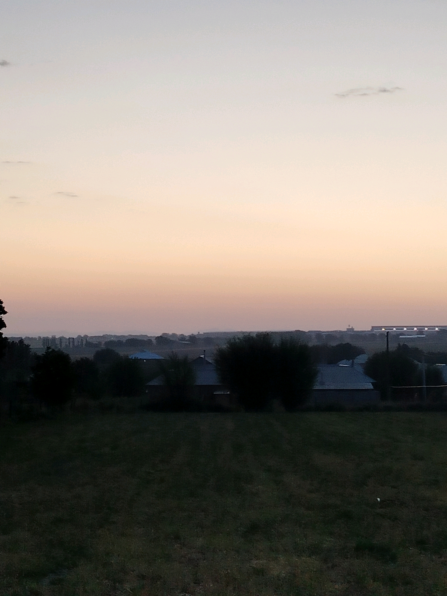 The image depicts a twilight scene over a rural or suburban area, likely in or near the city of Kölkent, Kazakhstan. The sky is a soft gradient of pale blue and peachy orange, suggesting either sunrise or sunset. A few wispy clouds are visible in the upper portion of the frame.
The middle ground shows a landscape with trees and the roofs of several buildings. Some of the buildings have dark, angular roofs, possibly made of metal or asphalt shingles. The vegetation is dense and dark, creating a silhouetted effect against the lighter sky. In the distance, a linear structure with multiple lights is visible, potentially a farm building, warehouse, or industrial facility.
The foreground is a field of dark grass, appearing somewhat dry and uneven. The overall atmosphere is quiet and still, characteristic of the transitional light of dawn or dusk. No people are visible in the image. The scene is devoid of any dynamic action. The environmental conditions suggest a clear but muted light.