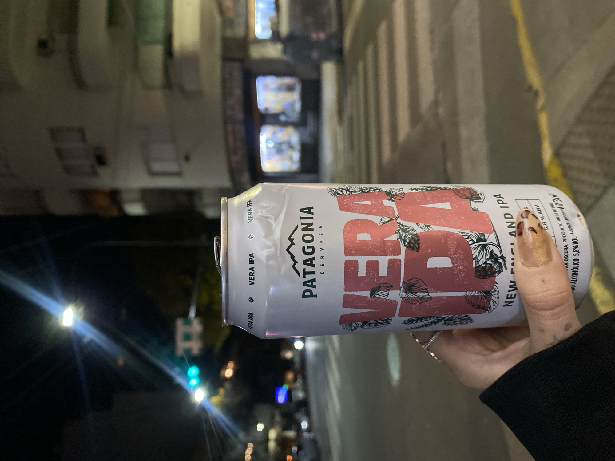 A person holds a can of Patagonia Vera IPA beer at night. The background is an urban street scene, possibly in Buenos Aires, Argentina, suggested by the beer brand. The can is white with red lettering and botanical illustrations, partially obscured by the hand. The street has buildings, streetlights, a crosswalk, and faint traffic signals. The person wears a black sleeve and has polished fingernails and rings.