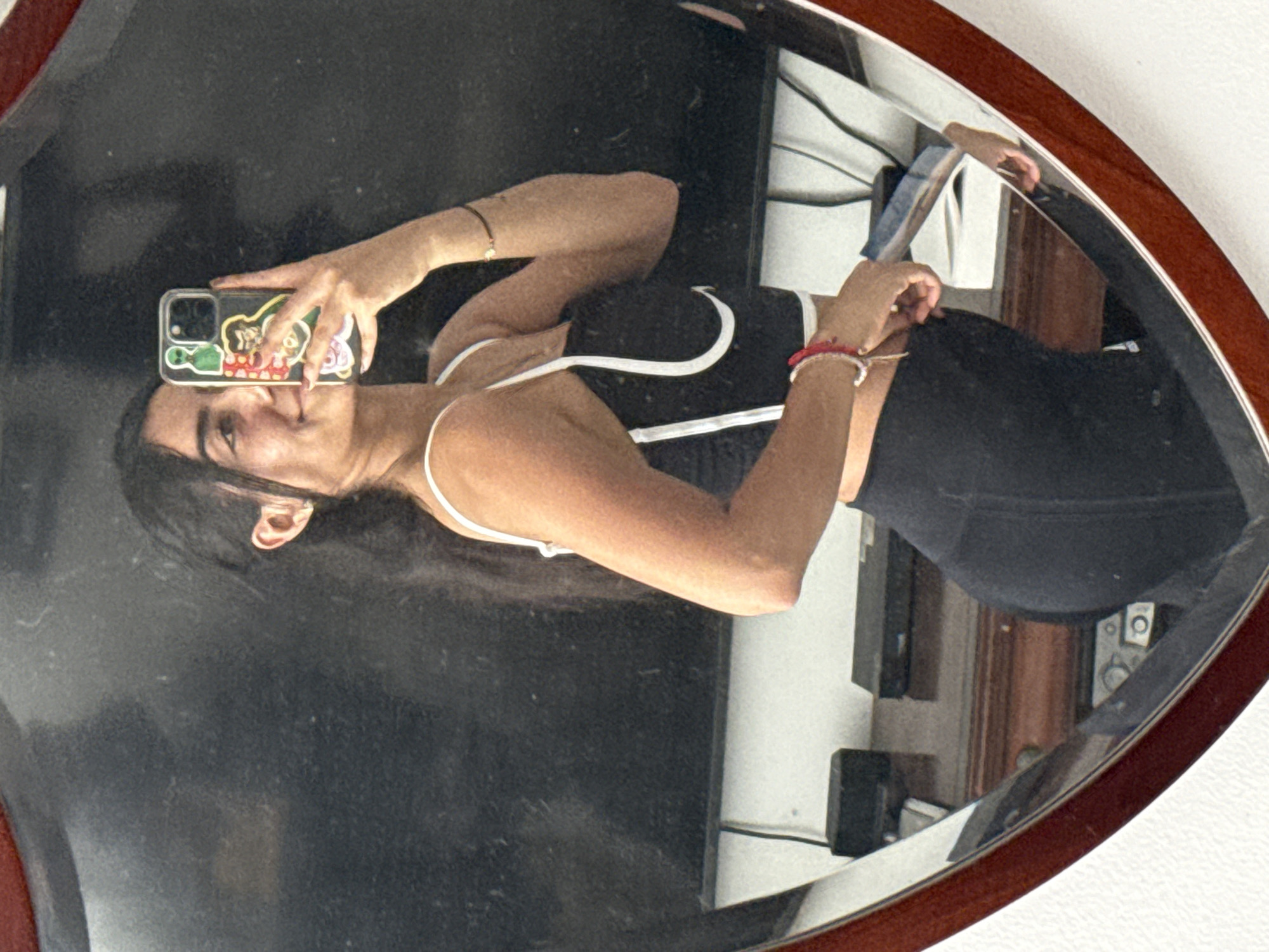 A woman is taking a selfie in a mirror. The mirror is heart-shaped with a brown wooden frame. The woman is wearing a black sports bra with white trim and black leggings. She is holding a cell phone in her right hand, and the phone case has several stickers on it. A silver bracelet is on her left wrist, and a red bracelet is on her right wrist. The background reflected in the mirror shows a dark-colored object which appears to be a TV screen. Also reflected in the mirror behind the woman is a partially visible room, with white walls, and a wooden doorframe. There is a white power strip with wires plugged into it. The location is Puebla, Mexico.