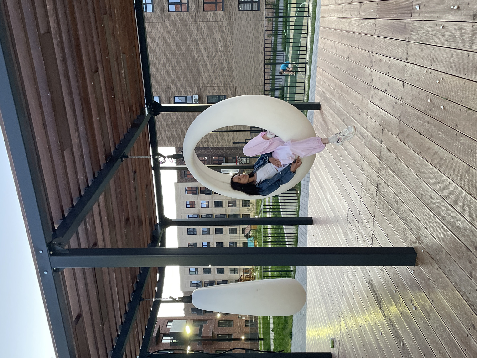 A young woman is sitting in a white, egg-shaped swing in a modern outdoor recreation area. The swing is suspended from a dark metal frame with a wooden slatted roof. The woman is wearing a white t-shirt, a denim jacket, and loose pink pants with white sneakers. She is looking down with a relaxed expression. To her left, another white, pear-shaped object is hanging from a similar swing mechanism. The area is paved with wooden planks, and a black metal railing encloses a green grassy area beyond. In the background, there are multi-story brick apartment buildings with numerous windows. One building is lit by exterior lights. Further back, a small playground structure and a basketball court are visible, with another person, possibly a child, on the court. The sky is clear and appears to be twilight. The overall scene suggests a newly developed residential complex with amenities for outdoor activities.