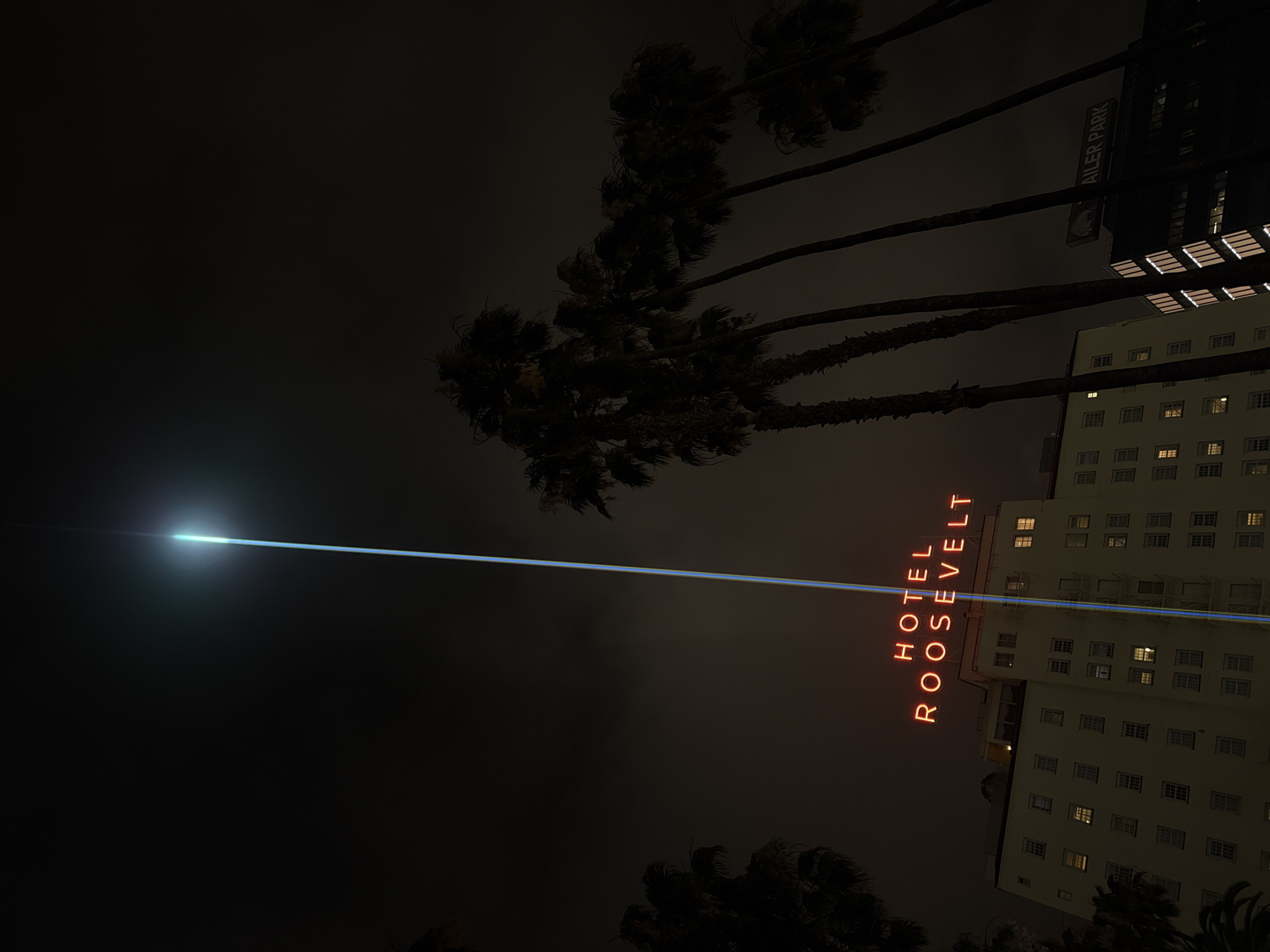 The image captures a night scene featuring the Roosevelt Hotel in Los Angeles, United States of America. The hotel is illuminated, and its vertical neon sign spelling out "HOTEL ROOSEVELT" is visible. Palm trees are silhouetted against the night sky. A bright light, possibly a spotlight or artificial light source, is positioned in the upper portion of the image. A sign for "Mueller Park" is also seen in the vicinity of the hotel. The overall environment is dark, suggesting it is nighttime.