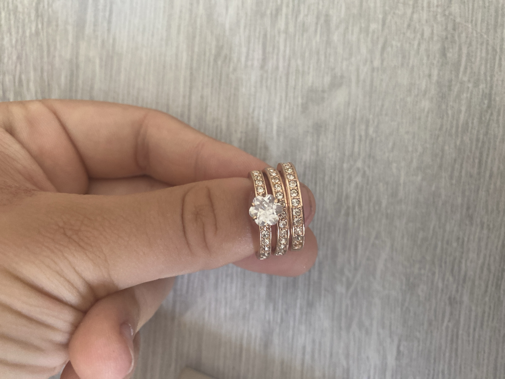 A close-up shot shows a person's hand holding a stack of three rose gold rings. The top ring features a prominent round-cut clear stone, likely a diamond or cubic zirconia, surrounded by smaller stones. The middle and bottom rings are bands encrusted with small, sparkling stones. The rings are stacked on one finger, suggesting they might be a wedding set or eternity bands. The background is a light-colored wooden surface with a visible grain pattern. The lighting is soft and natural, highlighting the sparkle of the stones and the metallic sheen of the rings. This image does not depict anything related to the city of Blagoevgrad, Bulgaria.