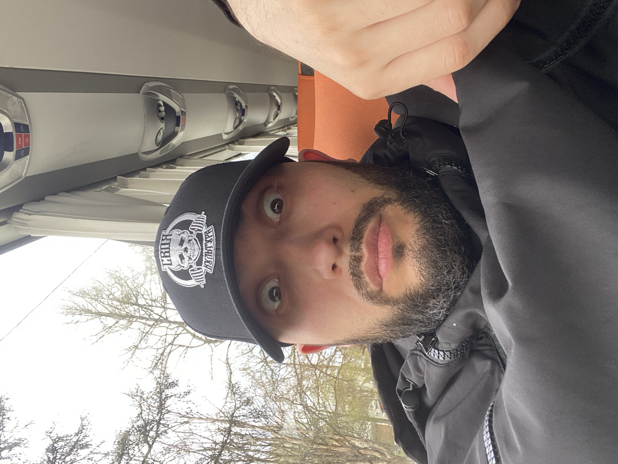 The image displays a selfie taken by a man inside what appears to be a public transport vehicle. The man, positioned centrally, has a dark beard and mustache and is looking directly at the camera with wide, neutral eyes. He wears a black baseball cap with a white logo featuring the text "TRUE BARBARY" (visible as "YABRAB EURT" due to mirroring) surrounding a skull graphic holding a barbell. He is also dressed in a black jacket. His right arm and hand are visible in the upper right portion of the frame.
The immediate background reveals the interior of the vehicle, characterized by light-colored overhead panels and multiple fixtures, possibly vents or reading lights. A red button labeled "STOP" is clearly visible on one of these panels, confirming the setting as a bus or train. Beyond the vehicle's window, an outdoor scene unfolds under an overcast sky, with numerous bare tree branches dominating the view. This suggests a cold season, likely late winter or early spring. The soft, diffused daylight indicates it is daytime. The scene captures a moment of travel in Opolie, Russia.