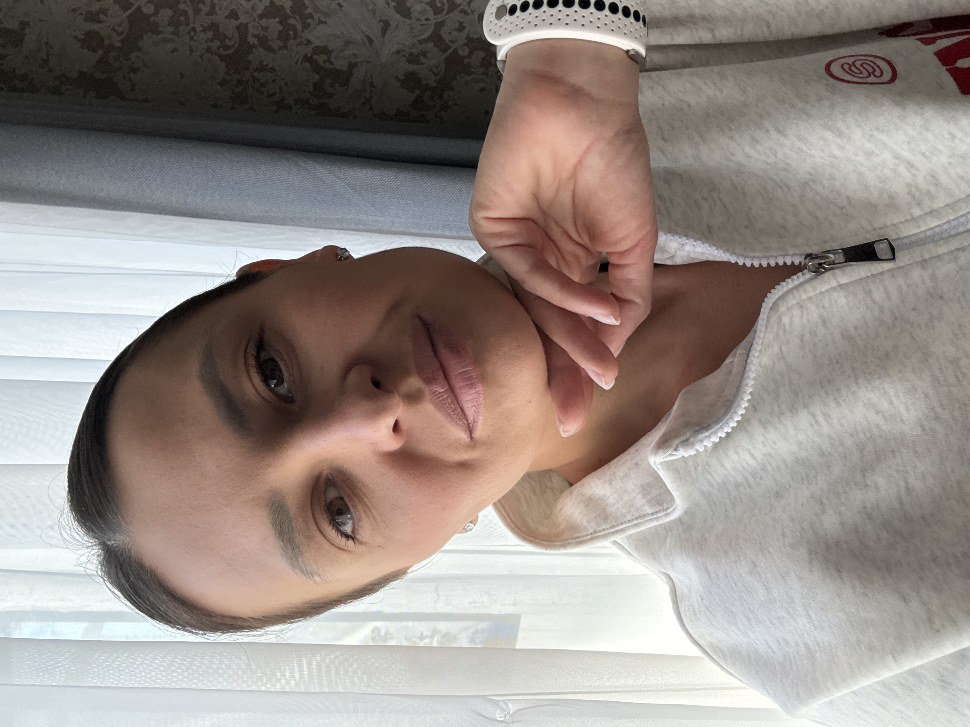 A woman is depicted from a low angle, facing the camera. She has brown hair pulled back, brown eyes, and light pink lipstick. Her right hand is raised to her neck. She wears a white smart watch with a black-dotted band on her wrist. She is wearing a white zippered sweatshirt. The background consists of white window blinds and a patterned curtain. There is no information in the image to confirm that this image was taken in the city of Chaikovsky, Russia.