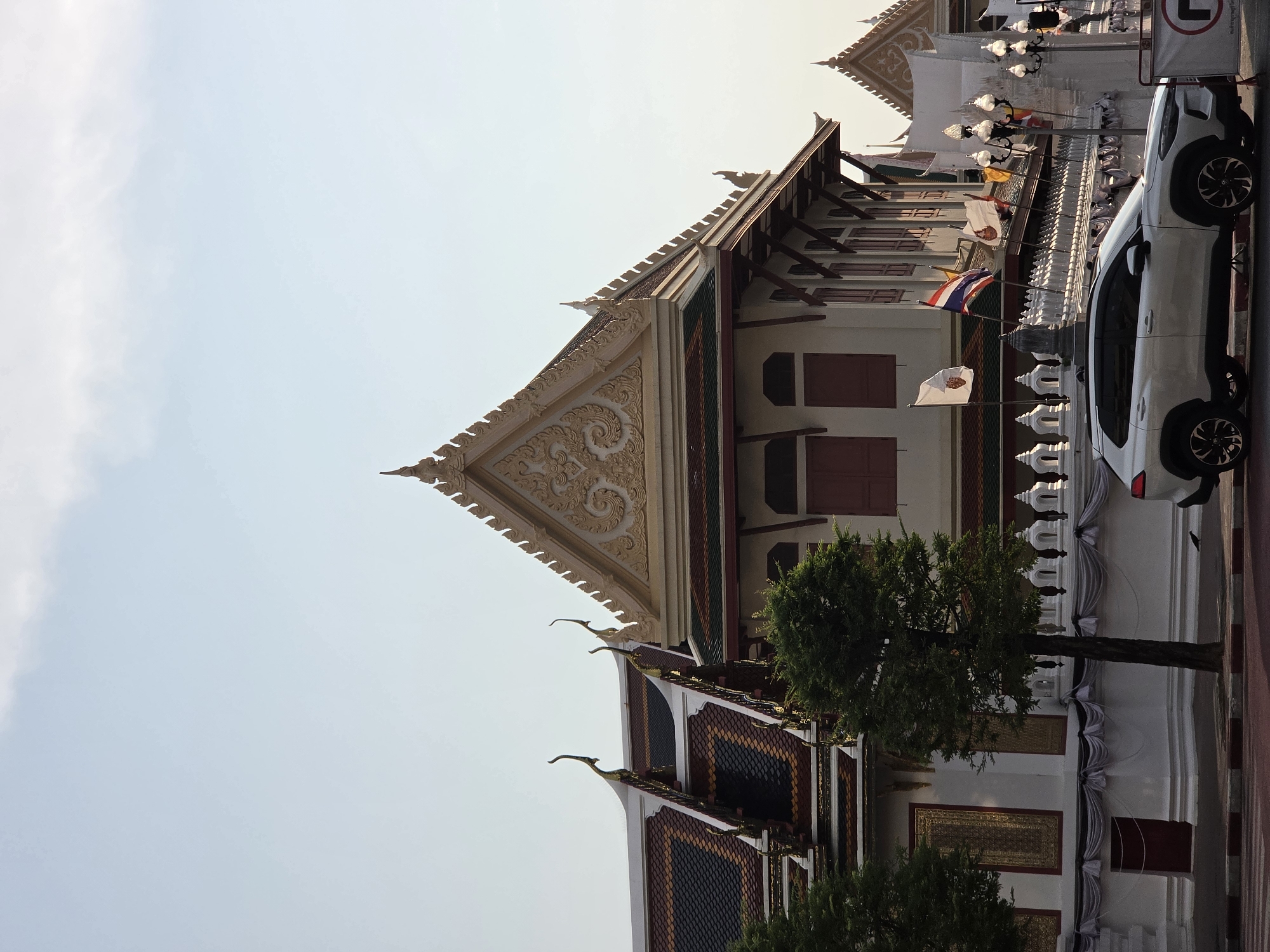 The image shows a section of a traditional Thai building, likely a temple or palace complex, in Bangkok, Thailand.
The building features intricate decorative elements on the roof and facades, including traditional patterns and ornate carvings. The roof is multi-tiered with red and blue tiles. The building's walls are white with windows covered by red shutters.
A white car is parked close to the building. Flags with a coat of arms and Thai national flags are mounted in front of the building. Two trees with green foliage are in the foreground.
The sky is light blue and clear.