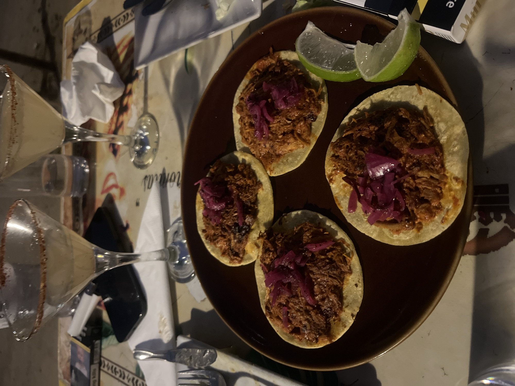 The image shows a plate of four tacos topped with meat and red cabbage, accompanied by two lime wedges, on a table. There are two cocktail glasses filled with drinks and rimmed with salt or sugar. A fork, knife, and napkins are also visible. The background includes a patterned tablecloth or placemat. Without additional information, it's impossible to confirm if the image was taken in Madrid, Spain.