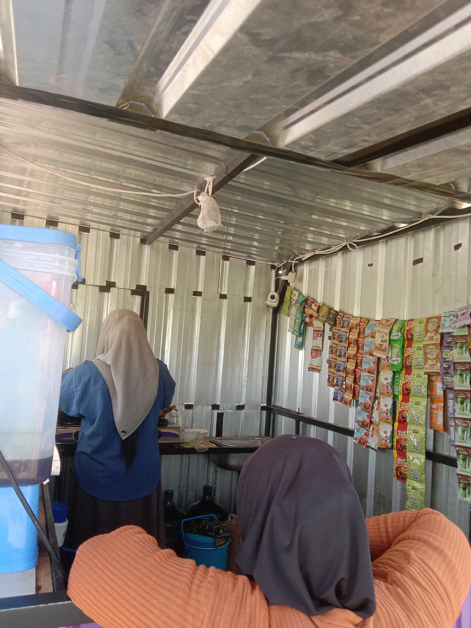 The image depicts an interior scene of what appears to be a small shop or food stall in Tulungrejo, Indonesia. The structure has a corrugated metal roof and walls. In the foreground, a person wearing an orange knitted top and a dark headscarf is visible from behind, seated. To their left, a large blue plastic container with a lid is present. In the middle ground, another person wearing a blue shirt and a light-colored headscarf is standing and facing away from the camera, appearing to be engaged in some activity at a counter or workspace. Behind this person, there is a sink and various containers. The back wall is lined with shelves holding numerous small packets of snacks or food items, some of which are hanging on display. Electrical wires and a light fixture are visible along the ceiling. The overall impression is of a functional, modest retail environment.