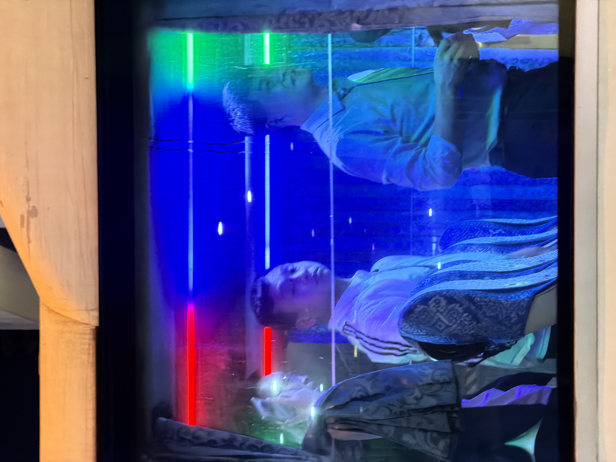 A vertical, indoor photograph depicts a television screen displaying a scene with two men. The screen features a modern aesthetic with illuminated red, blue, and green vertical lines. One man, dressed in a light-colored shirt and dark pants, appears to be floating near the top of the screen. Below him, another man is lying down and covered with a blue patterned blanket. The entire scene seems to be reflecting within the television screen, creating a mirrored, layered effect. The television is encased in a wooden frame, partially visible on the left and right sides of the image. The image does not provide any information to determine the location in the city of Đà Nẵng, Vietnam.